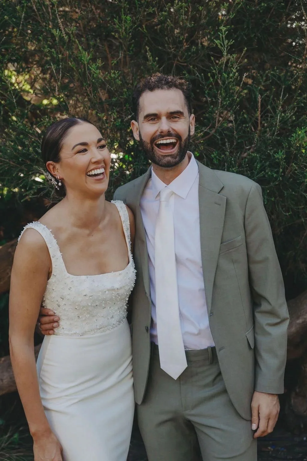 A newlywed couple smiling outdoors, the bride in a white dress with beaded details and the groom in a light gray suit, standing in front of leafy greenery.