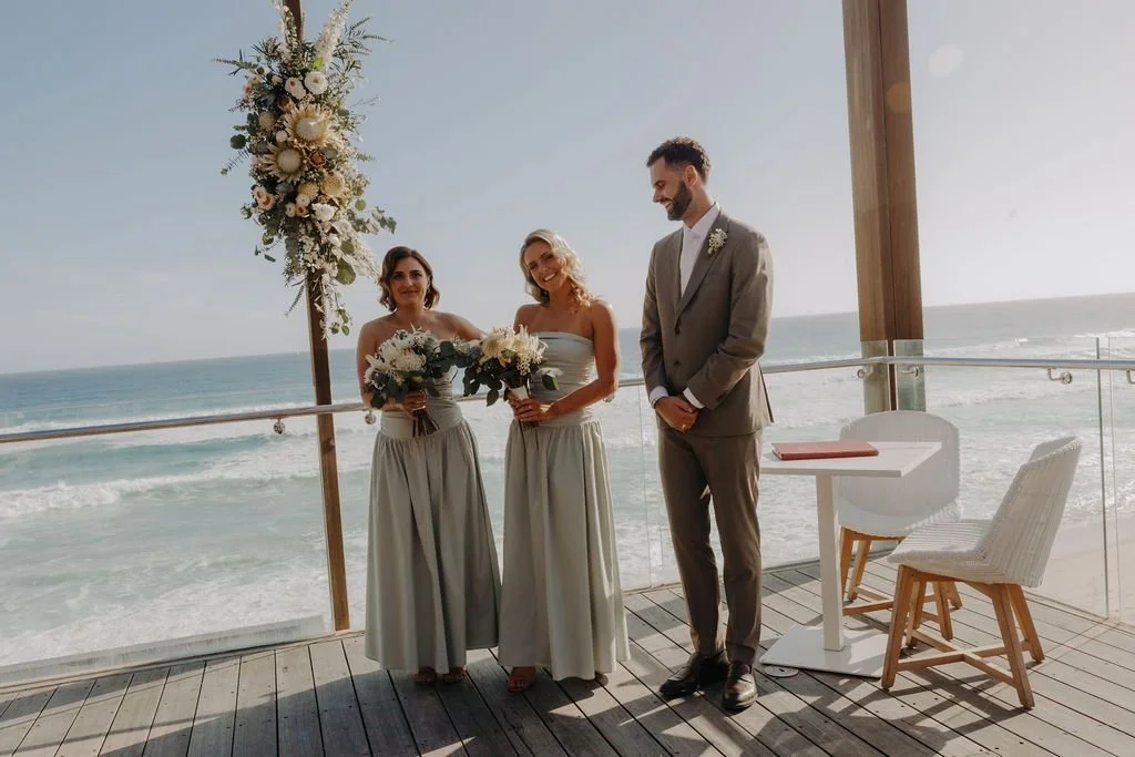 A wedding ceremony taking place on an outdoor deck overlooking the beach, with two women in matching dresses holding bouquets and a man in a suit standing beside them, under a floral arch.