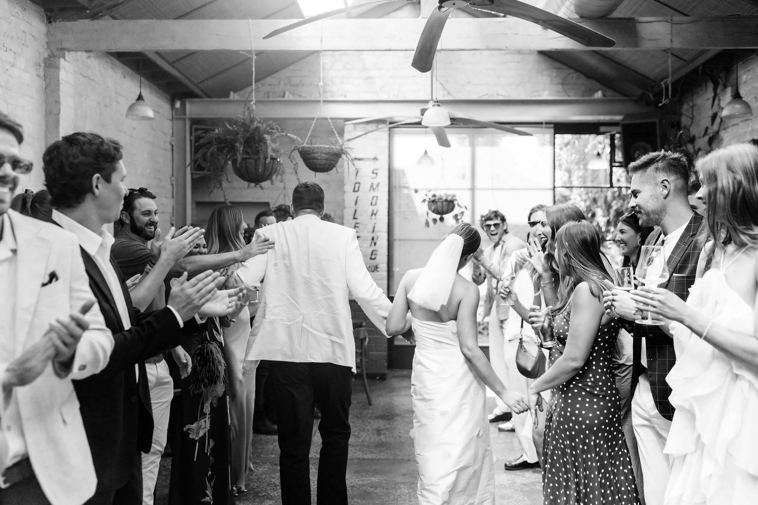 Group of people at a wedding reception, standing in a circle, clapping and smiling, with the bride and groom in the center surrounded by friends and family indoors with hanging plants and large windows.