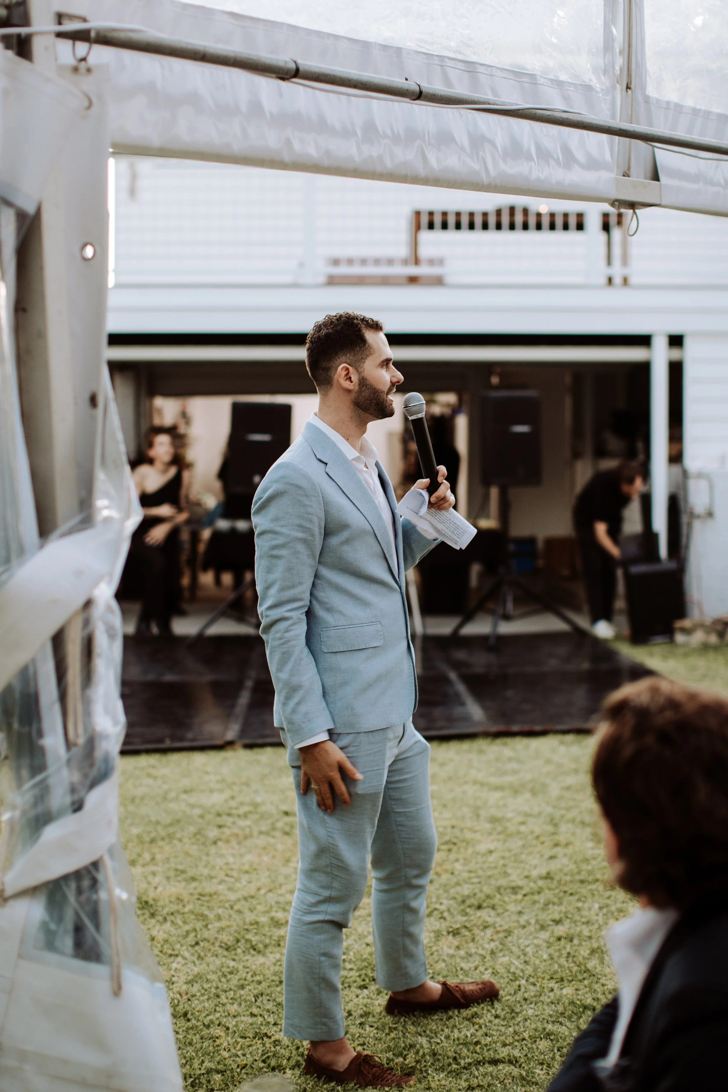 A man in a light gray suit and brown loafers speaking into a microphone during an outdoor event on a grassy area, with some people and speakers in the background.
