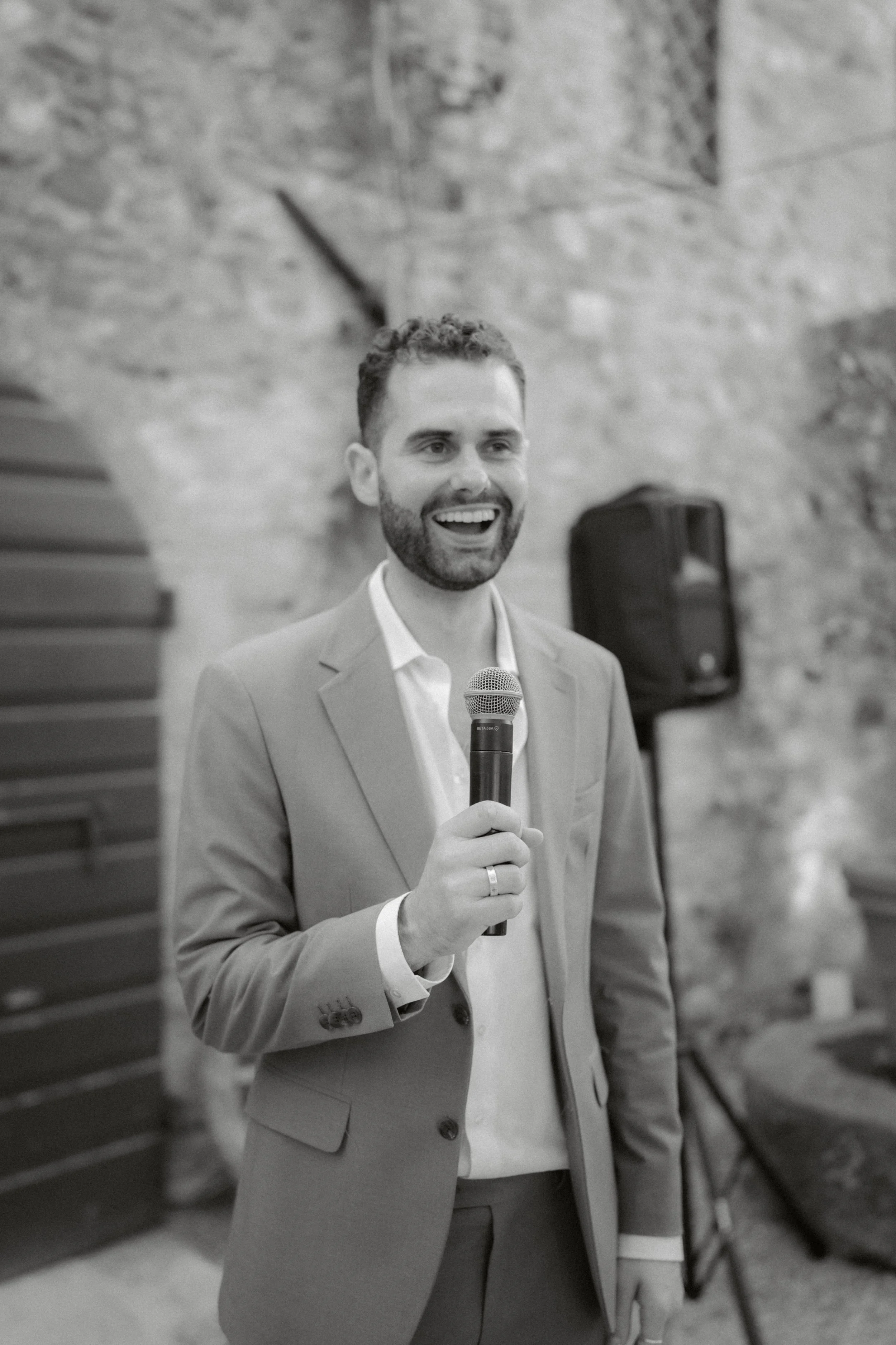 A man with short curly hair and a beard, wearing a light-colored suit, is smiling and speaking into a microphone. He is indoors with a stone wall background and a speaker in the background.