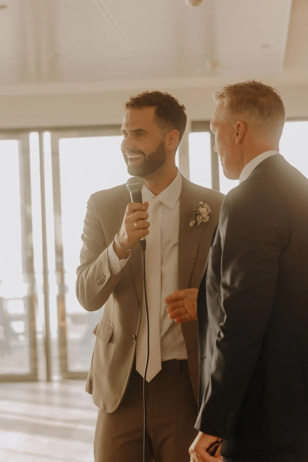 Two men speaking at a wedding reception, one holding a microphone, both dressed in suits, with a bright window in the background.