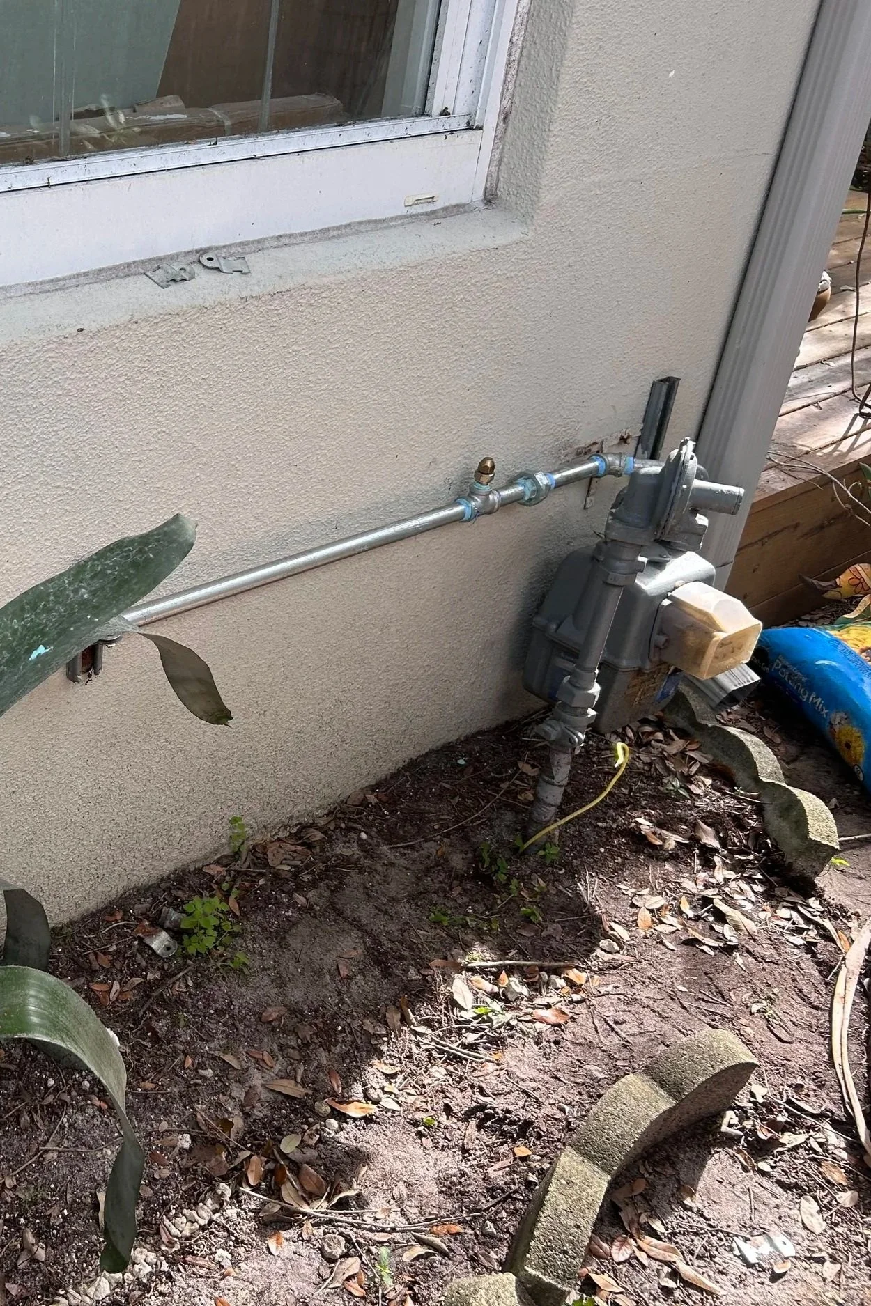 Outdoor view of a house wall with a natural gas meter and piping, surrounded by dirt, leaves, and a partially built stone border.