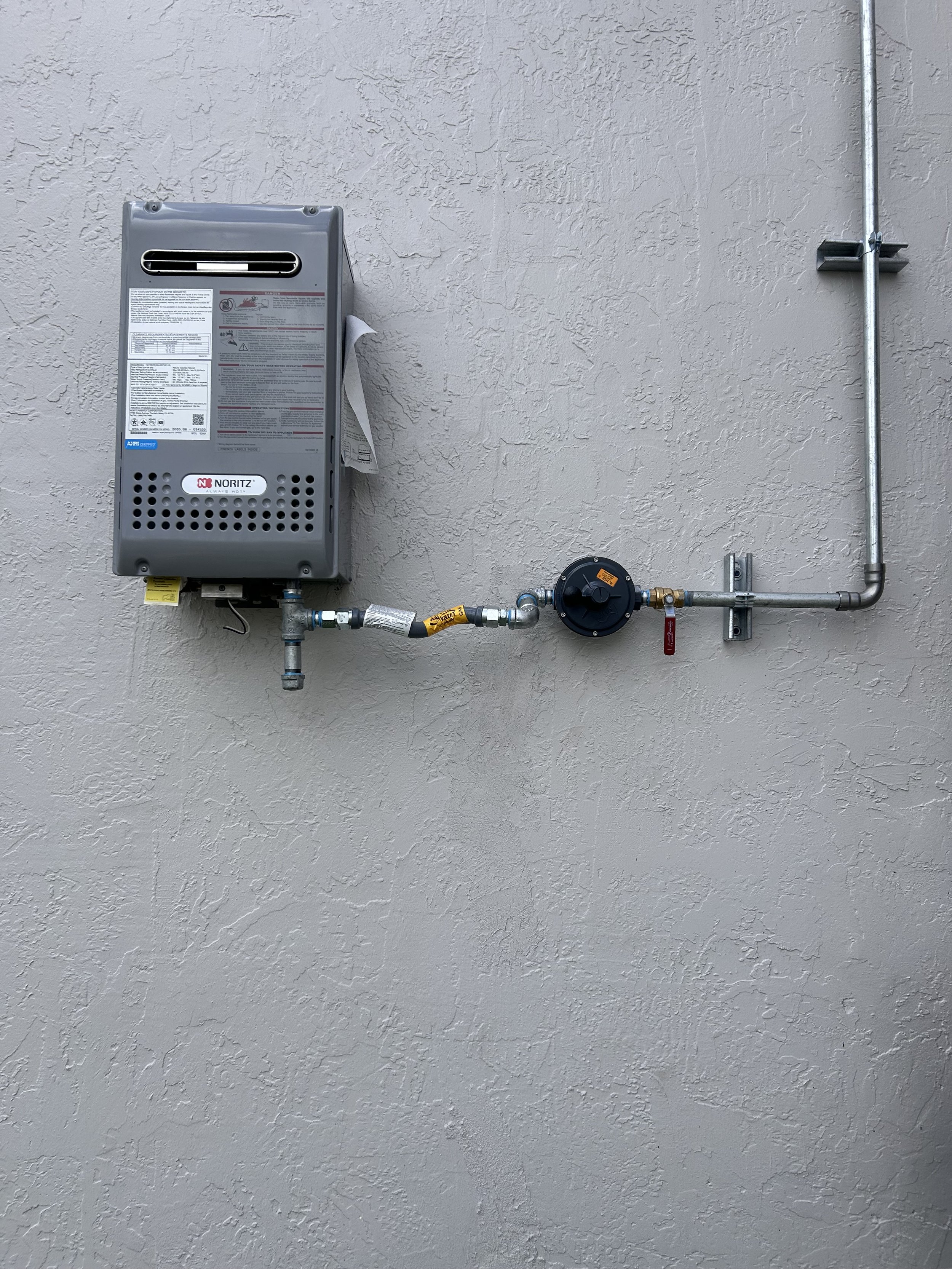 A wall-mounted gas water heater with pipes, valves, and a control panel, installed on a stucco-textured beige wall.