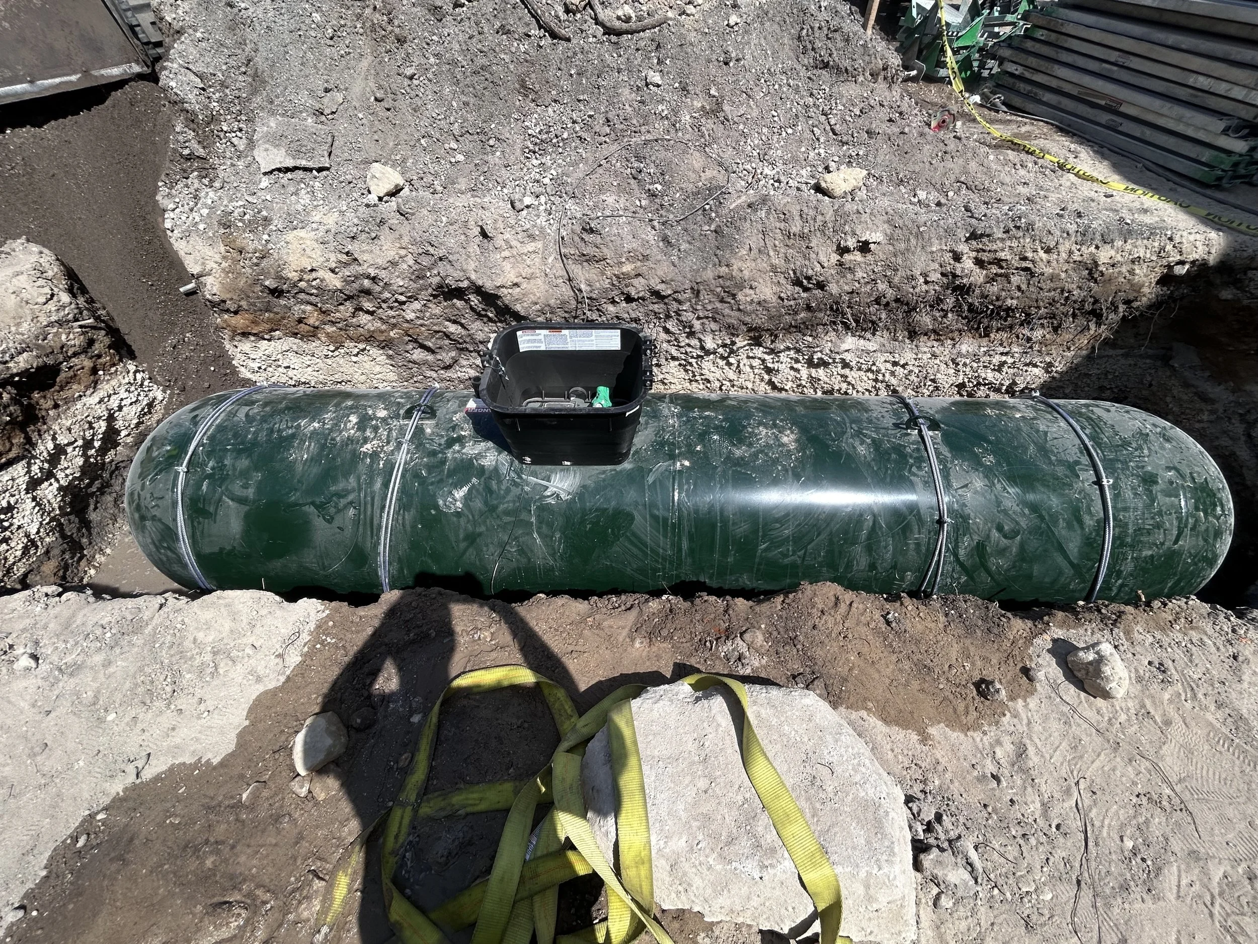 Close-up view of construction site with a large pipe laid underground, surrounded by dirt, rocks, and construction equipment, including a yellow strap and a black toolbox on top of the pipe.