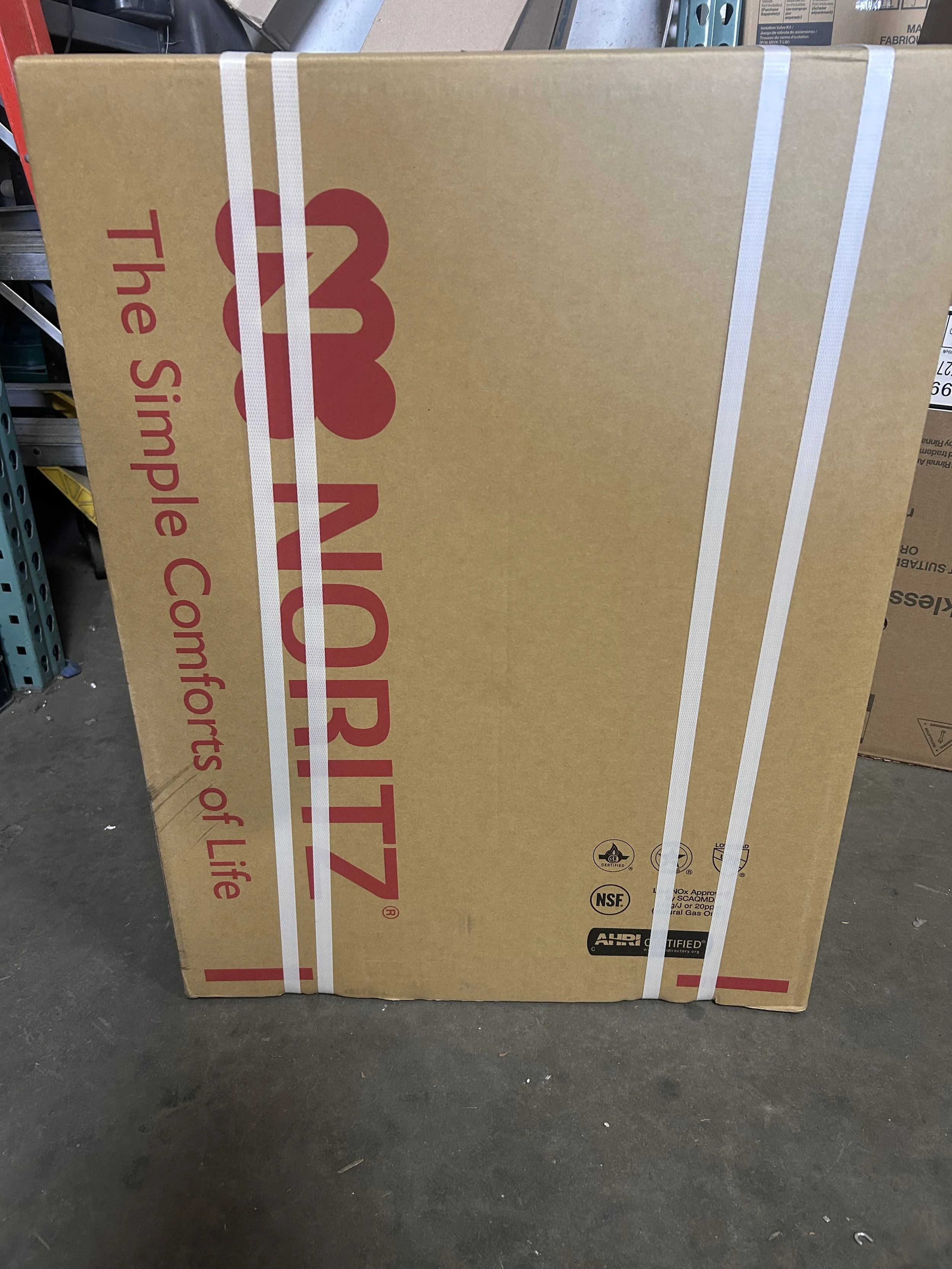 A large cardboard box labeled 'The_IMPORTZ' with additional text 'The Simple Comforts of Life,' secured with white straps, standing on a concrete floor in a warehouse or storage area.
