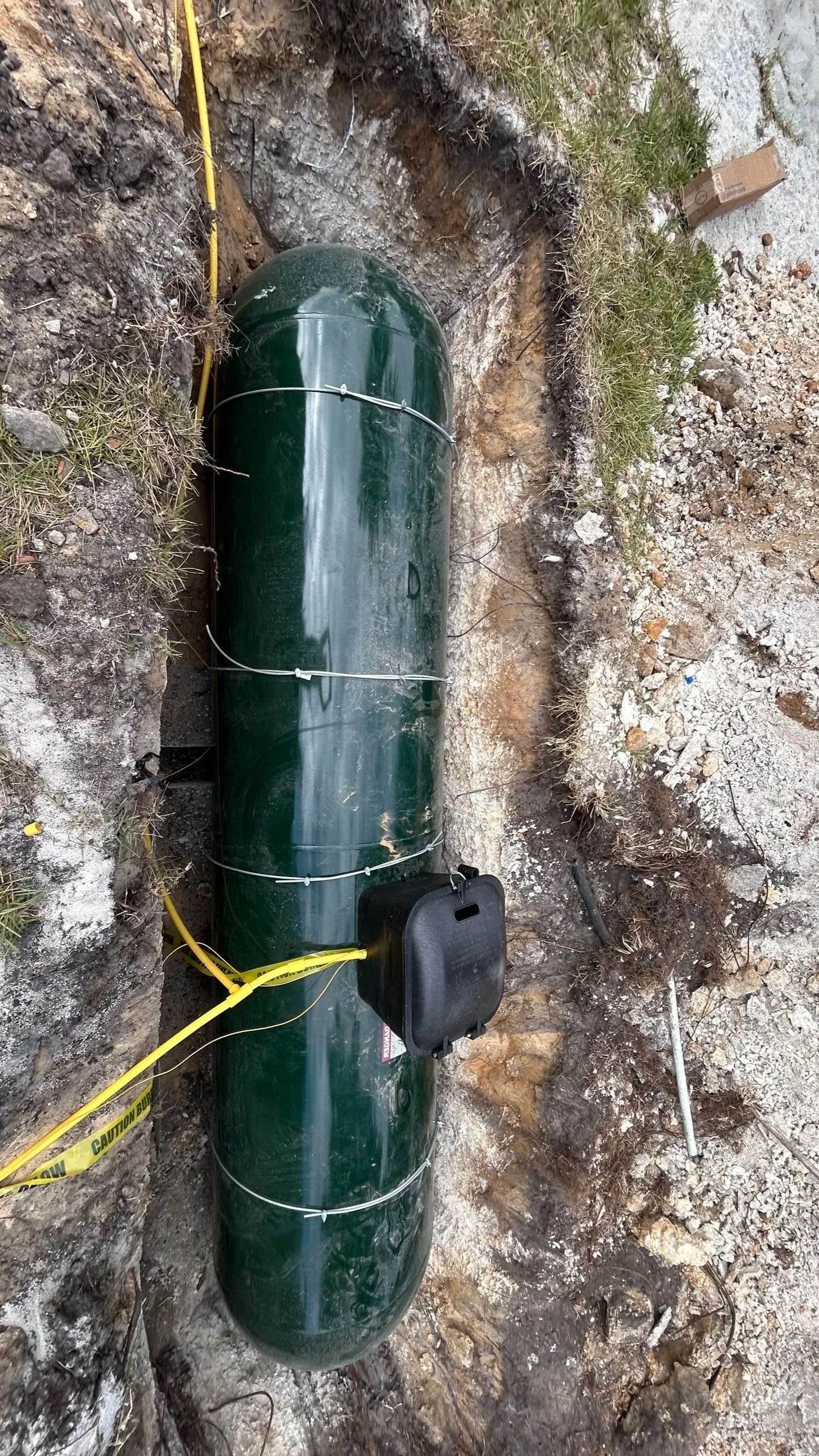 Underground 1000Gal Propane tank with black protective dome, underground piping, and equipment inside a dirt area with gravel and grass.