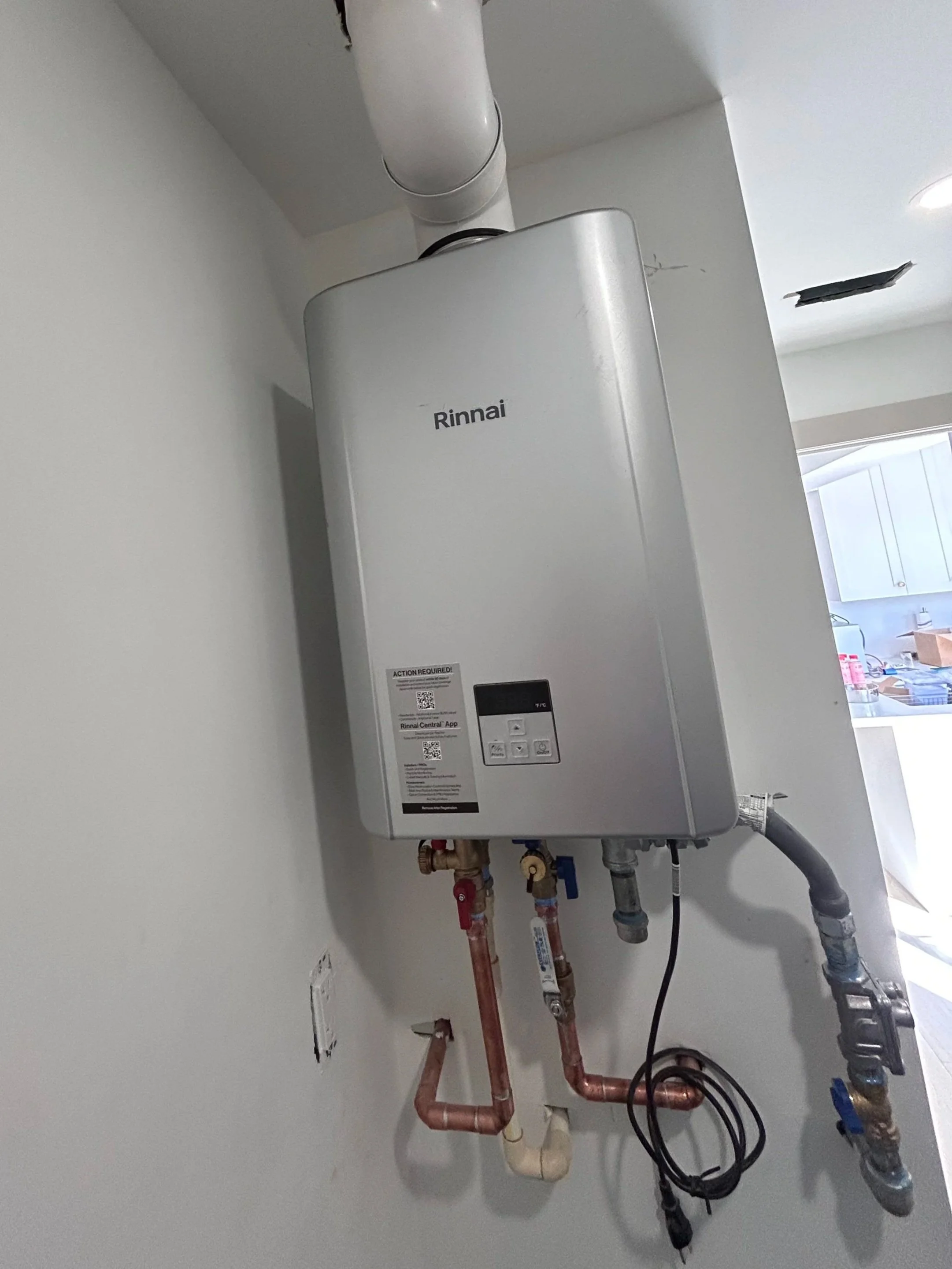 An indoor wall-mounted Rinnai water heater with connected pipes and hoses, situated near a white wall and ceiling, with an open kitchen visible in the background.