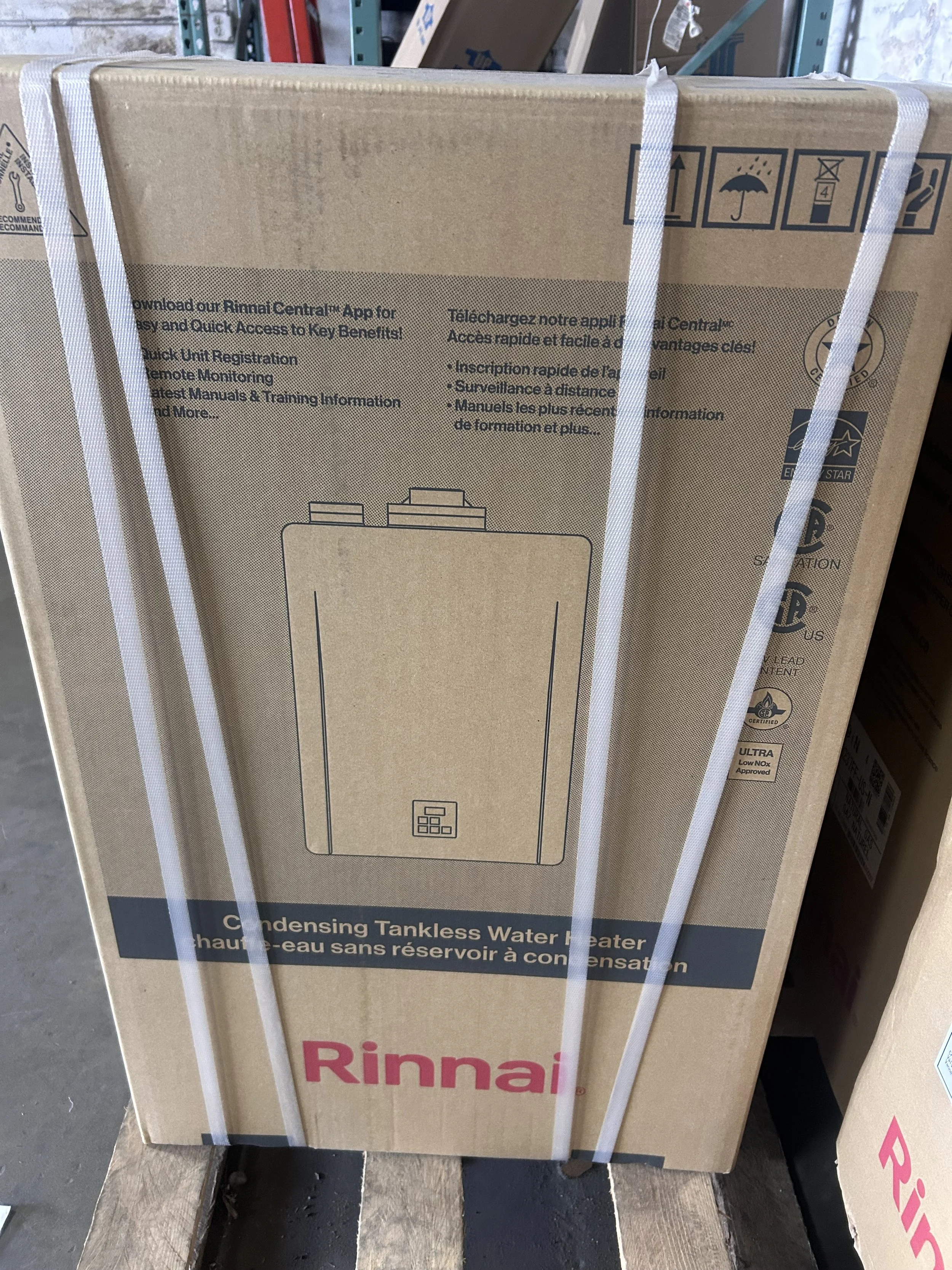 Boxed Rinnai Tankless Water Heater with printed diagram and text, wrapped with white straps, placed on a wooden pallet in a warehouse.