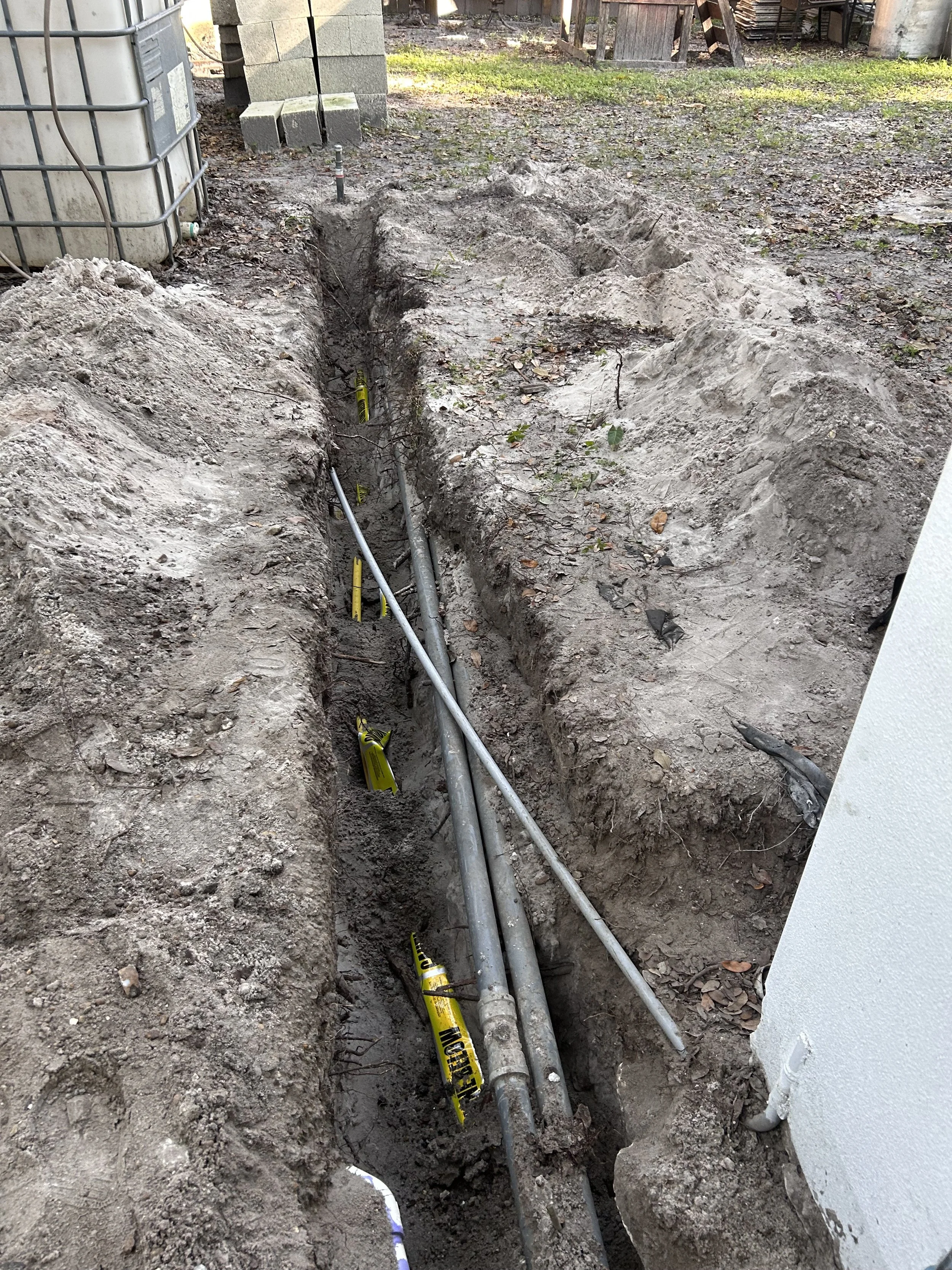 Underground utility excavation with pipes and cables, marked with yellow warning tape, beside a white building, outdoor area with dirt, gravel, and construction materials in the background.
