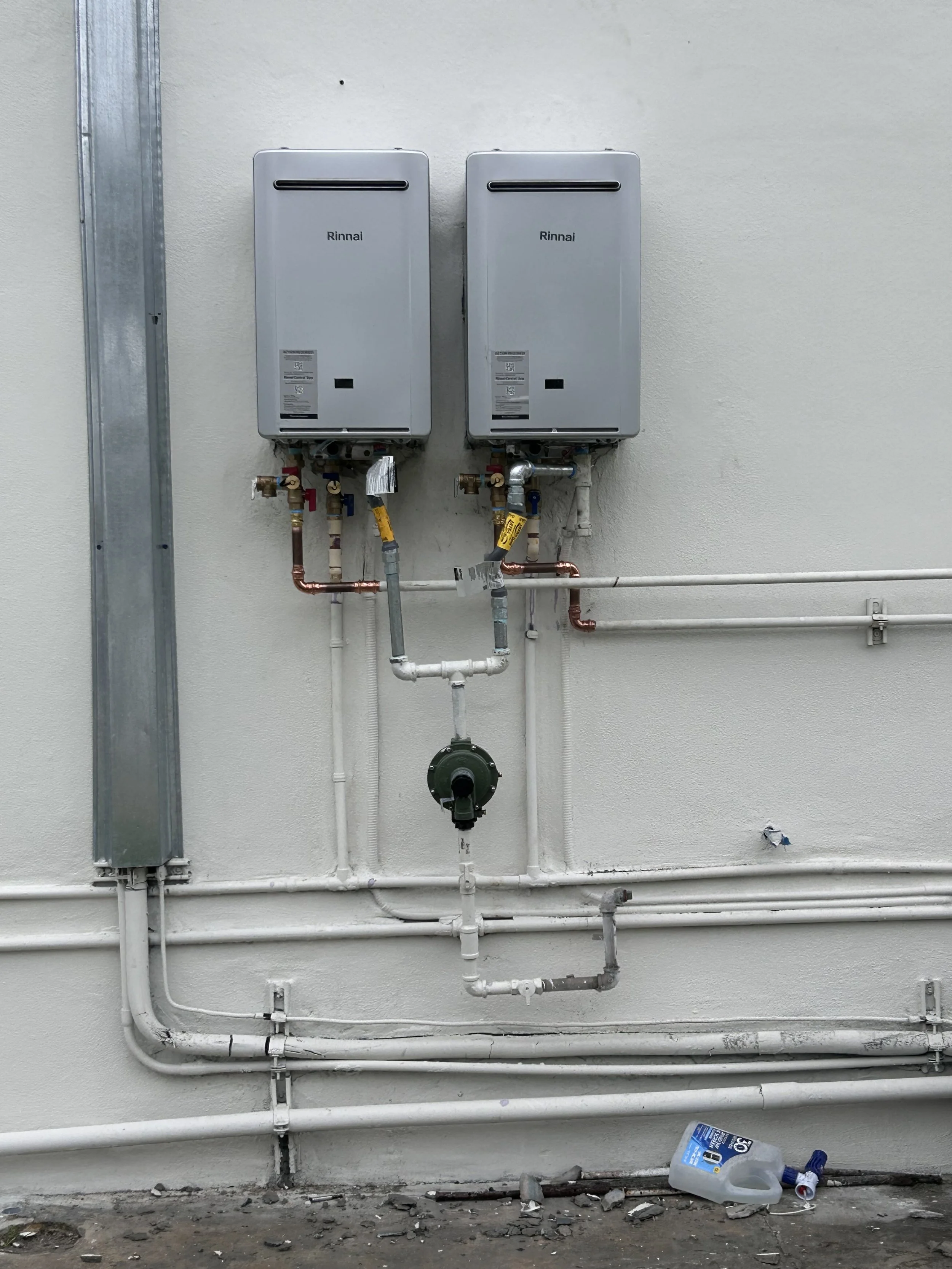 Two Rinnai tankless water heaters installed on a white wall with pipes and gauges underneath, and a gallon jug of cleaning solution on the ground.