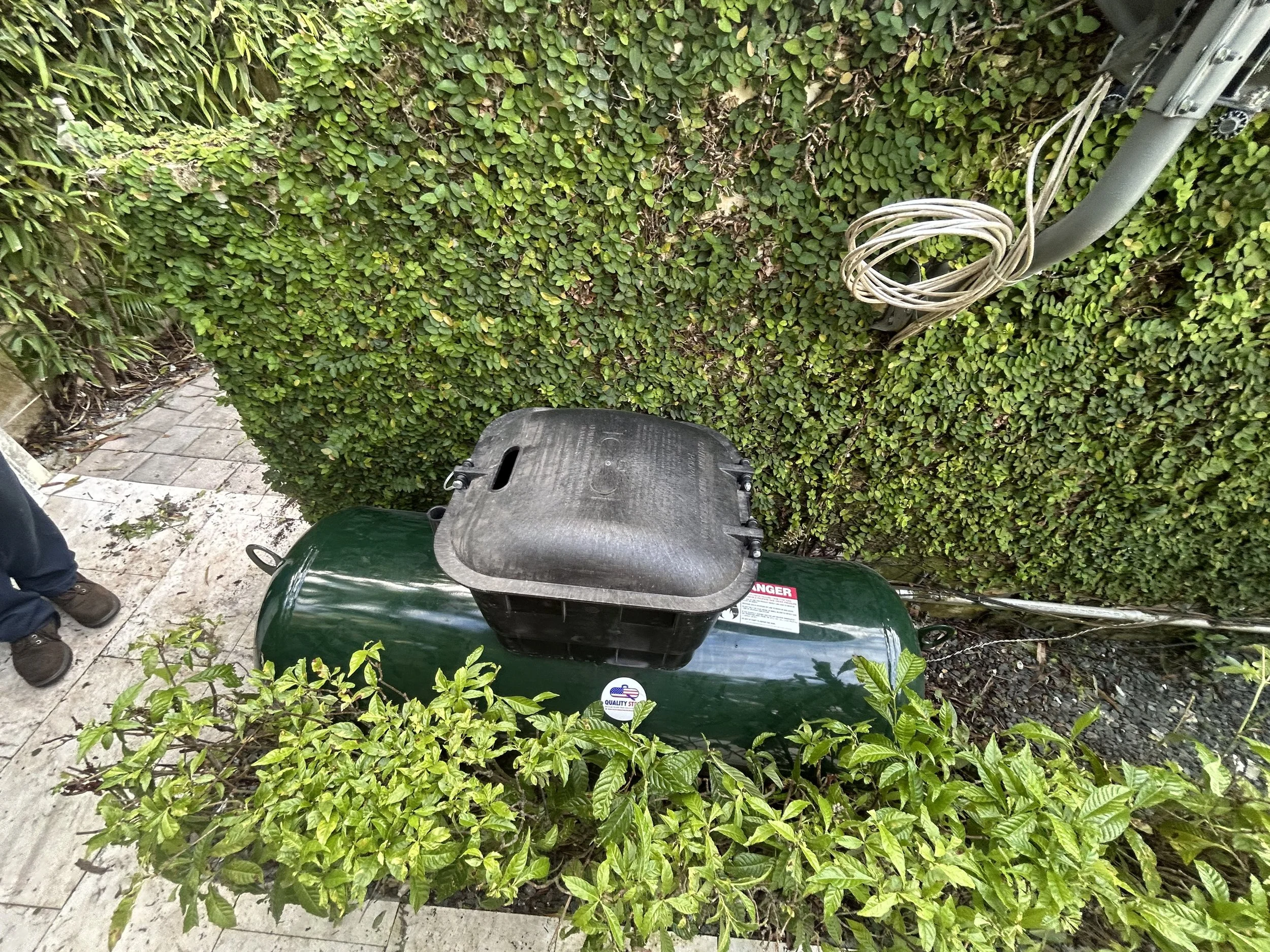 Green cylindrical propane tank with black regulator and hose connected, situated outdoors next to a hedge and brick pathway.
