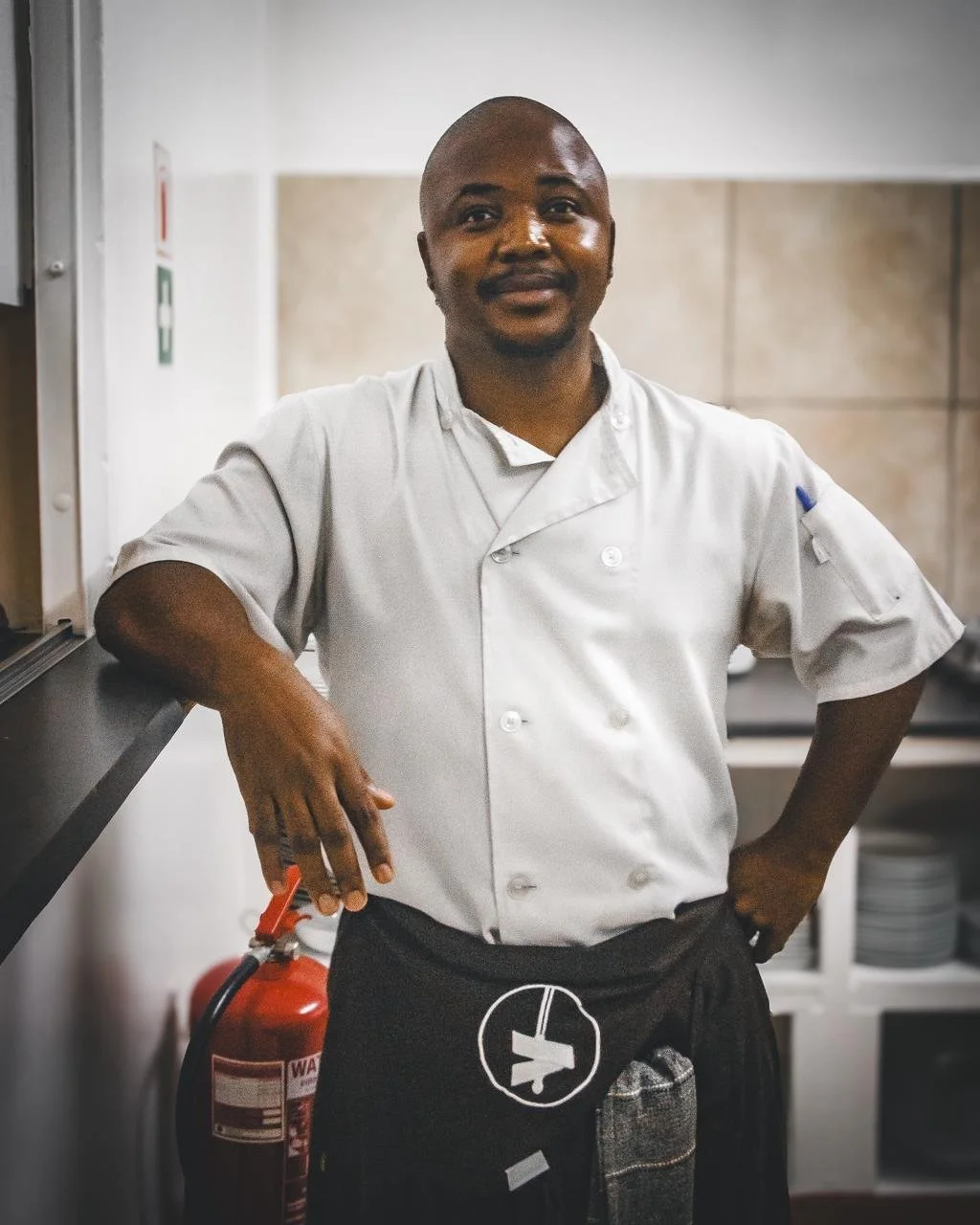 A male chef in a white uniform stands in a kitchen, smiling, with his right arm on a counter and his left hand on his hip.