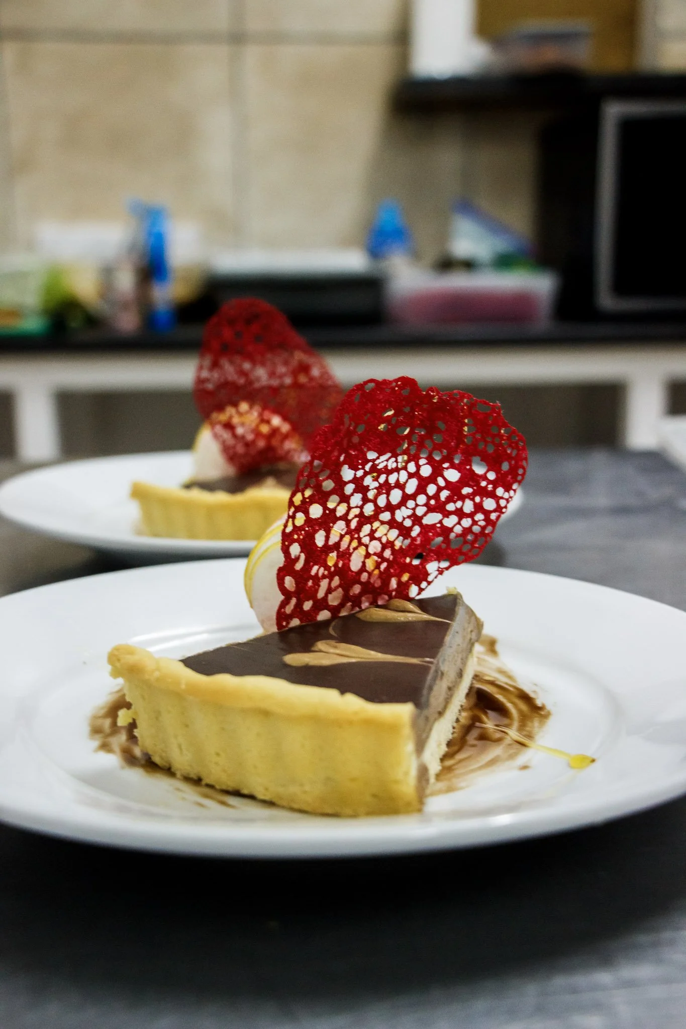 Two plates with slices of chocolate and vanilla layered cheesecake topped with red decorative sugar lace and cream on a dark surface, with a blurred kitchen background.