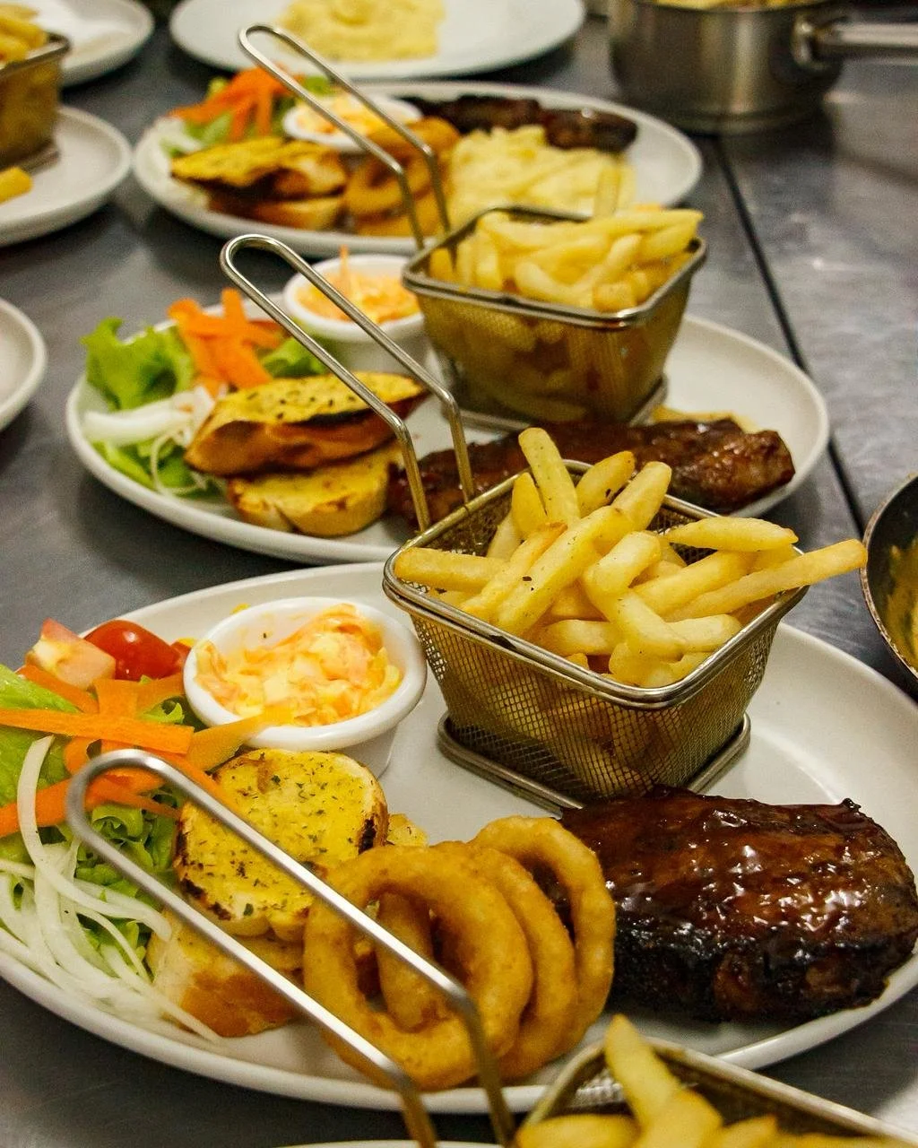 Multiple plates of food on a table, featuring French fries, onion rings, grilled meat, salad, and dipping sauces.