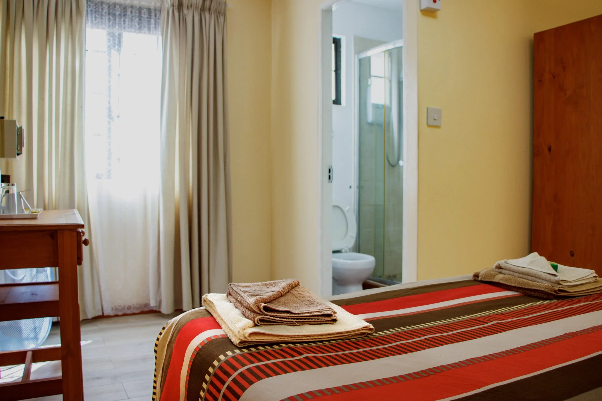 Hotel room with a bed covered in a striped red, brown, and beige bedspread, towels on the bed, a small wooden table with a tray and kettle, beige curtains, and an open door to a bathroom with a toilet and shower.