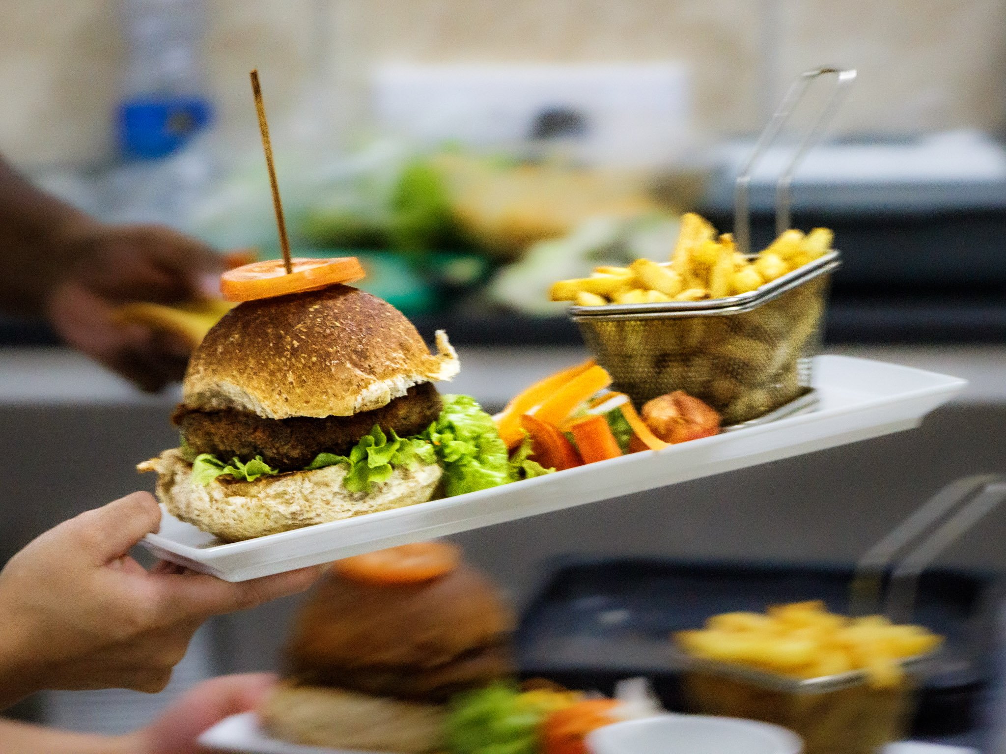 A person holding a white tray with a burger, a small portion of French fries in a metal basket, and some sliced vegetables. The burger has lettuce and a patty with a bun and a slice of tomato on top, held together with a skewer. The background shows a counter with food and kitchen equipment.