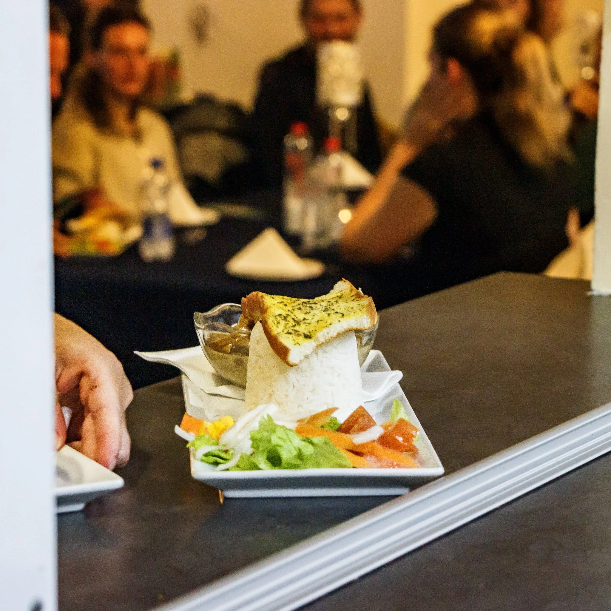 A plate of food with rice, grilled chicken, salad, and a bread slice with herb butter, set on a table in front of blurred group of people at a dinner event.