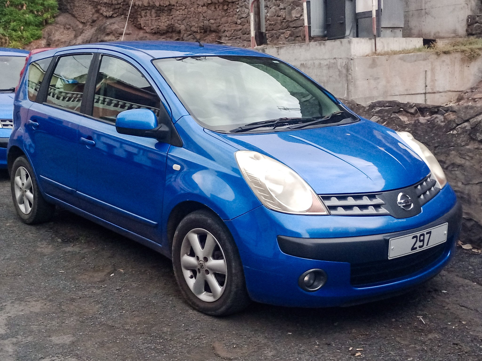 Blue Nissan hatchback parked on a dirt surface with a rocky and concrete background.