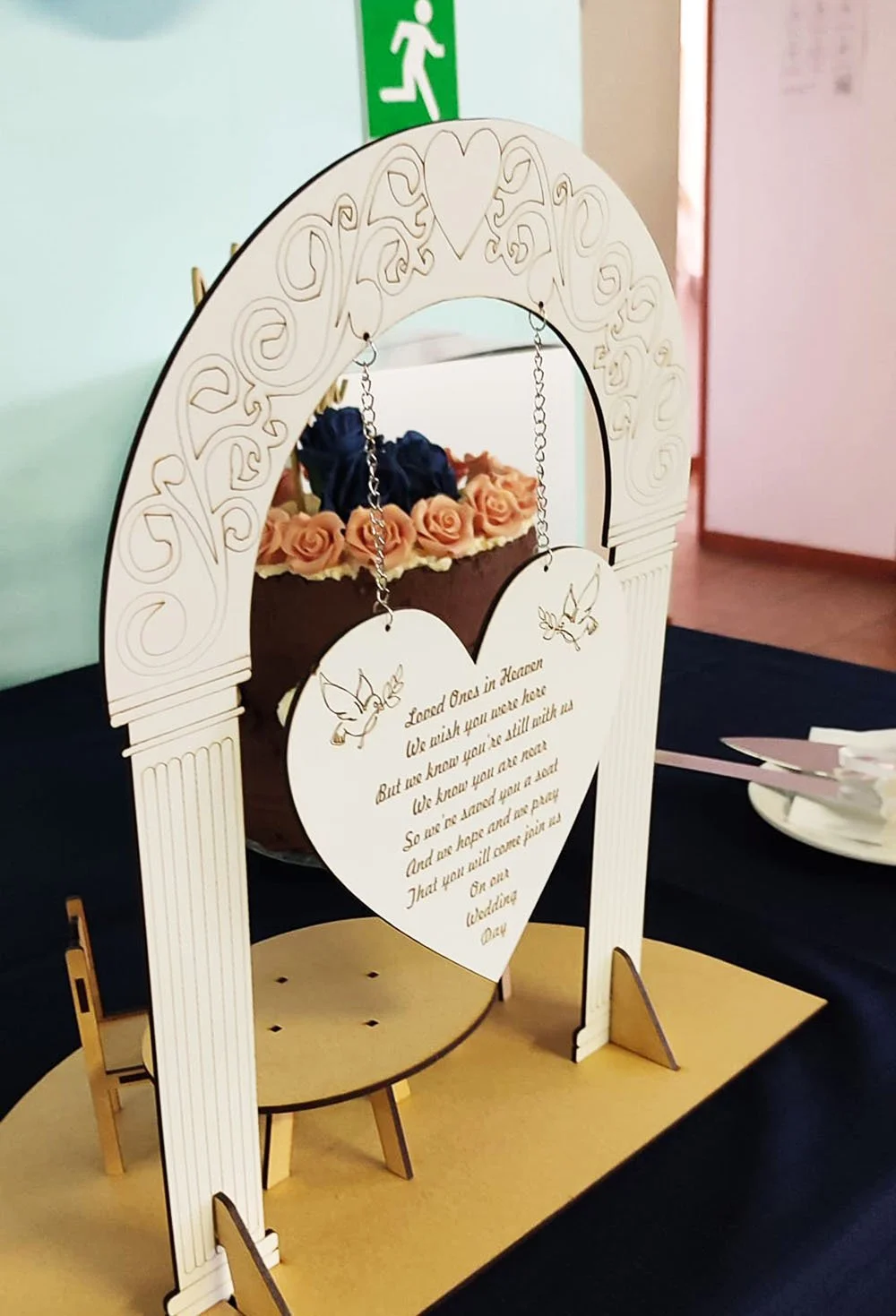 A decorative wedding or romantic event sign with a poem, featuring two engraved angel illustrations, surrounded by a white ornate arch and a heart-shaped cutout, placed on a table with flowers and cutlery in the background.