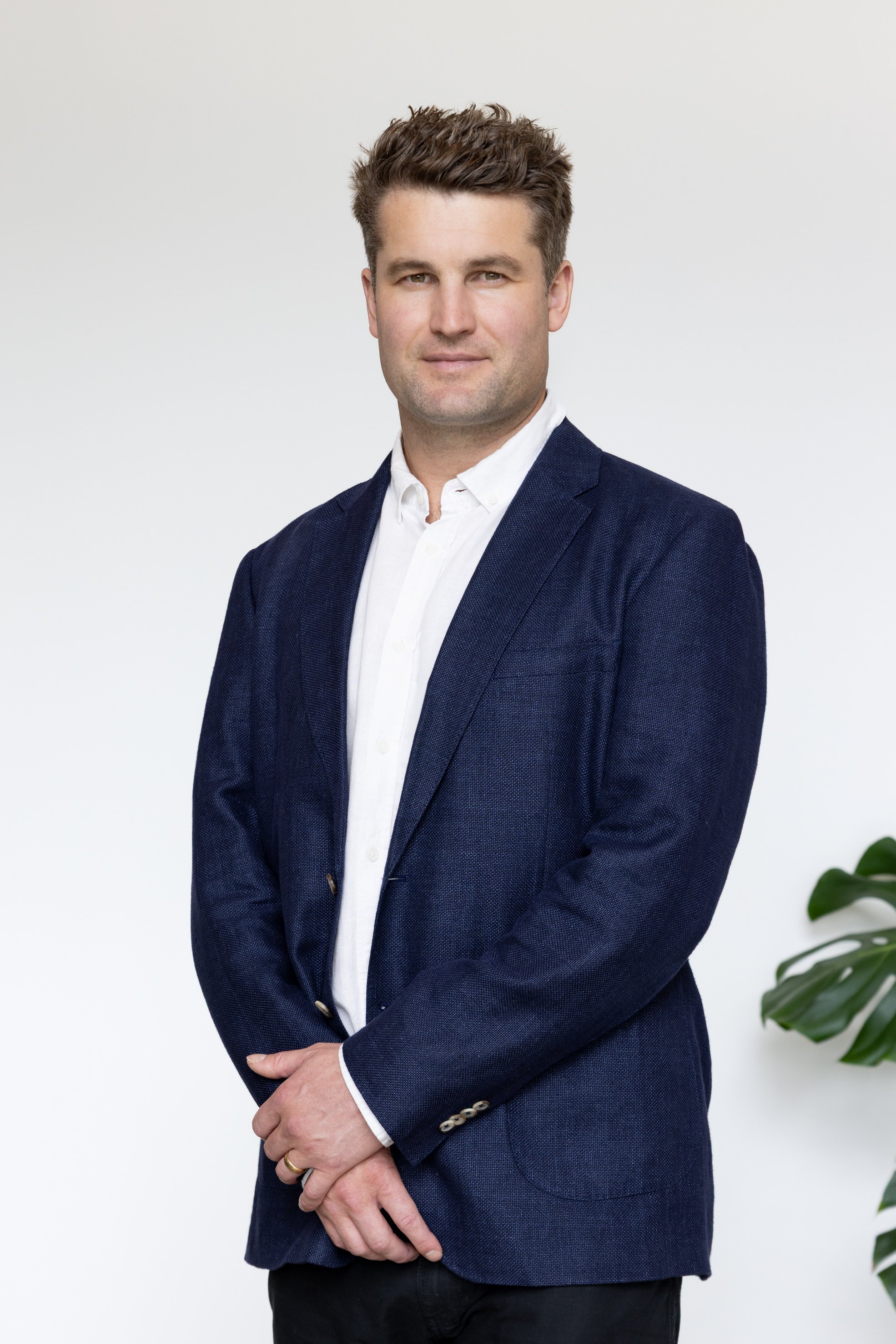 A man wearing a dark blue blazer and white shirt standing against a plain white wall, with a plant partially visible on the right side.