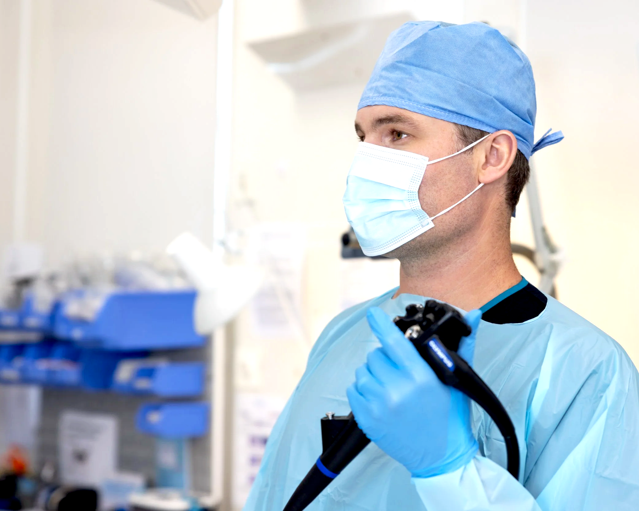 A male healthcare worker wearing blue scrubs, a surgical mask, and a blue surgical cap, working in a medical facility or hospital, performing an endoscopic procedure