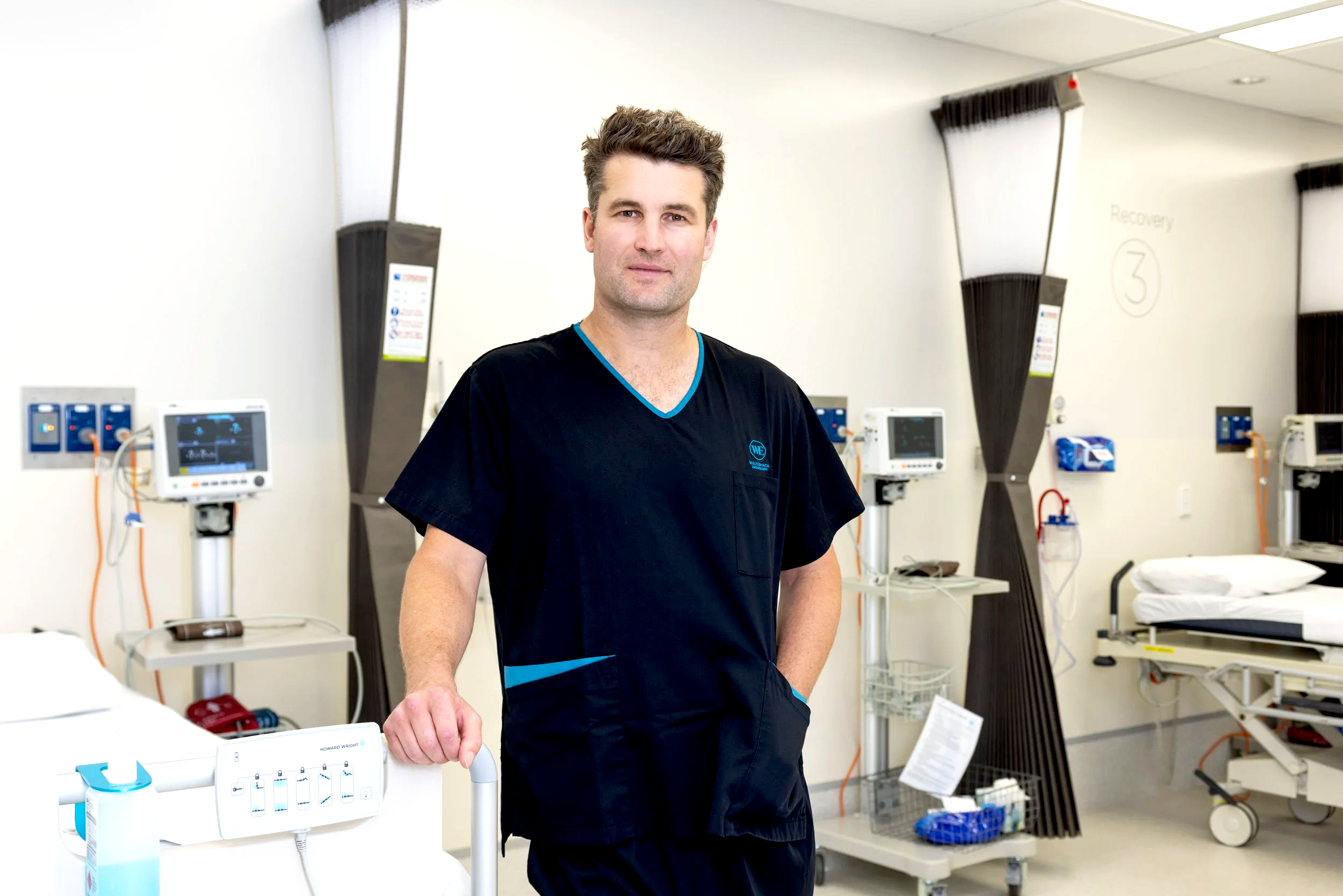 A male healthcare professional standing in a hospital room with medical equipment and monitors in the background.
