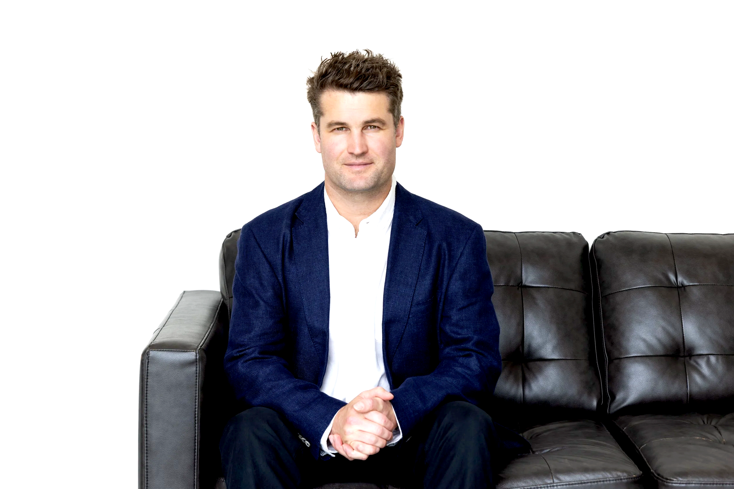 A man in a white shirt and navy blazer sitting on a black leather couch, with hands clasped, smiling slightly, against a plain white background.