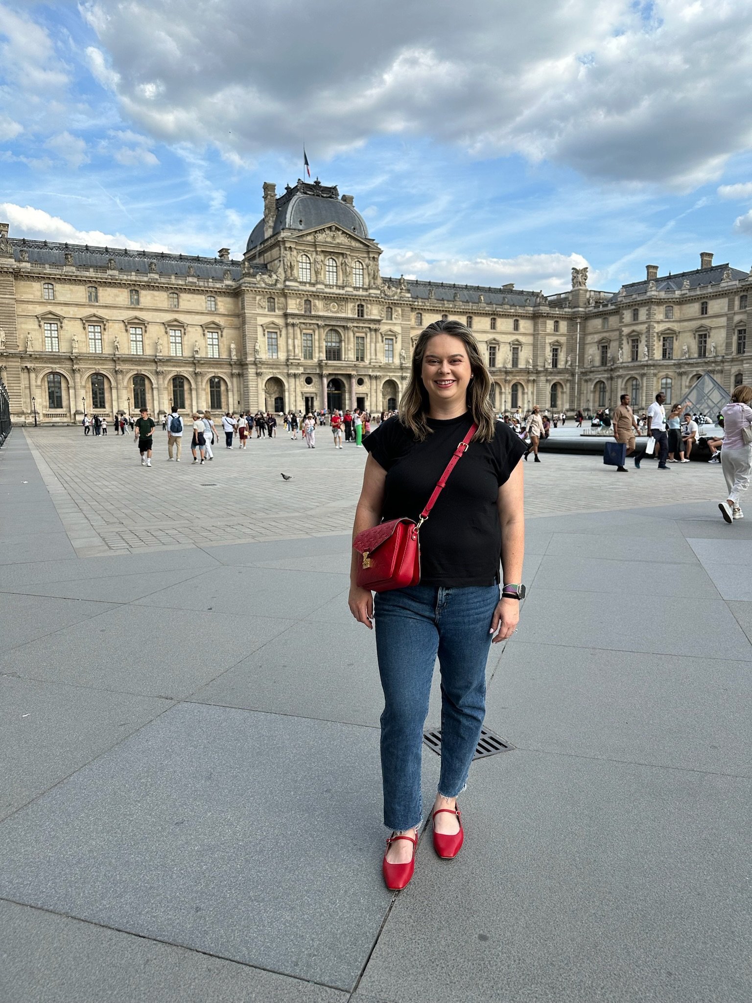 A woman in black top, blue jeans, and red shoes standing in front of a historic building, with a red purse across her shoulder, smiling at the camera.
