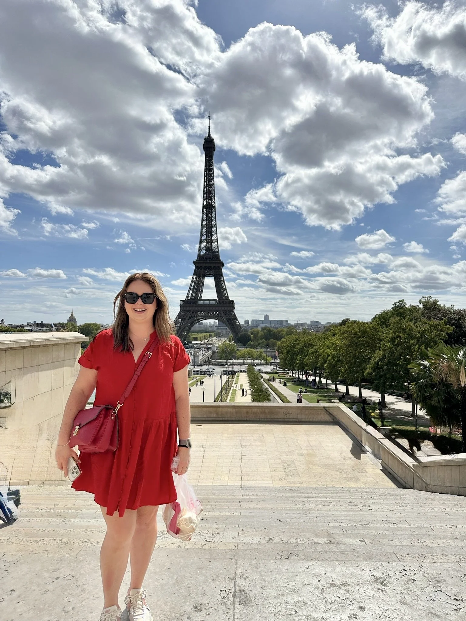 A woman in a red dress and sunglasses standing on stairs with the Eiffel Tower in the background during daytime with scattered clouds.