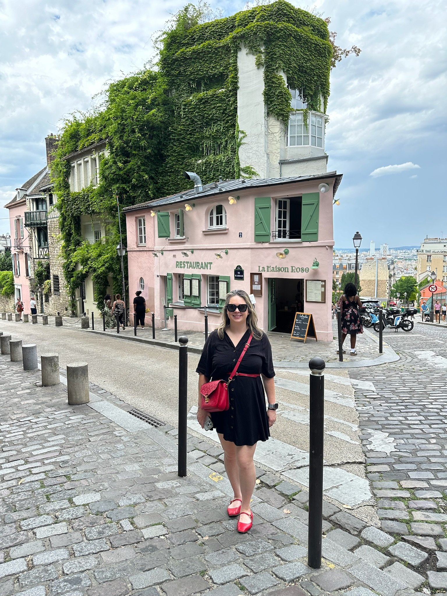 A woman wearing a black dress and red shoes standing on a cobblestone street with a pink building behind her that has green shutters, with several other people walking nearby. The building has grass and vines on the upper levels and is located in a European city, likely Paris.