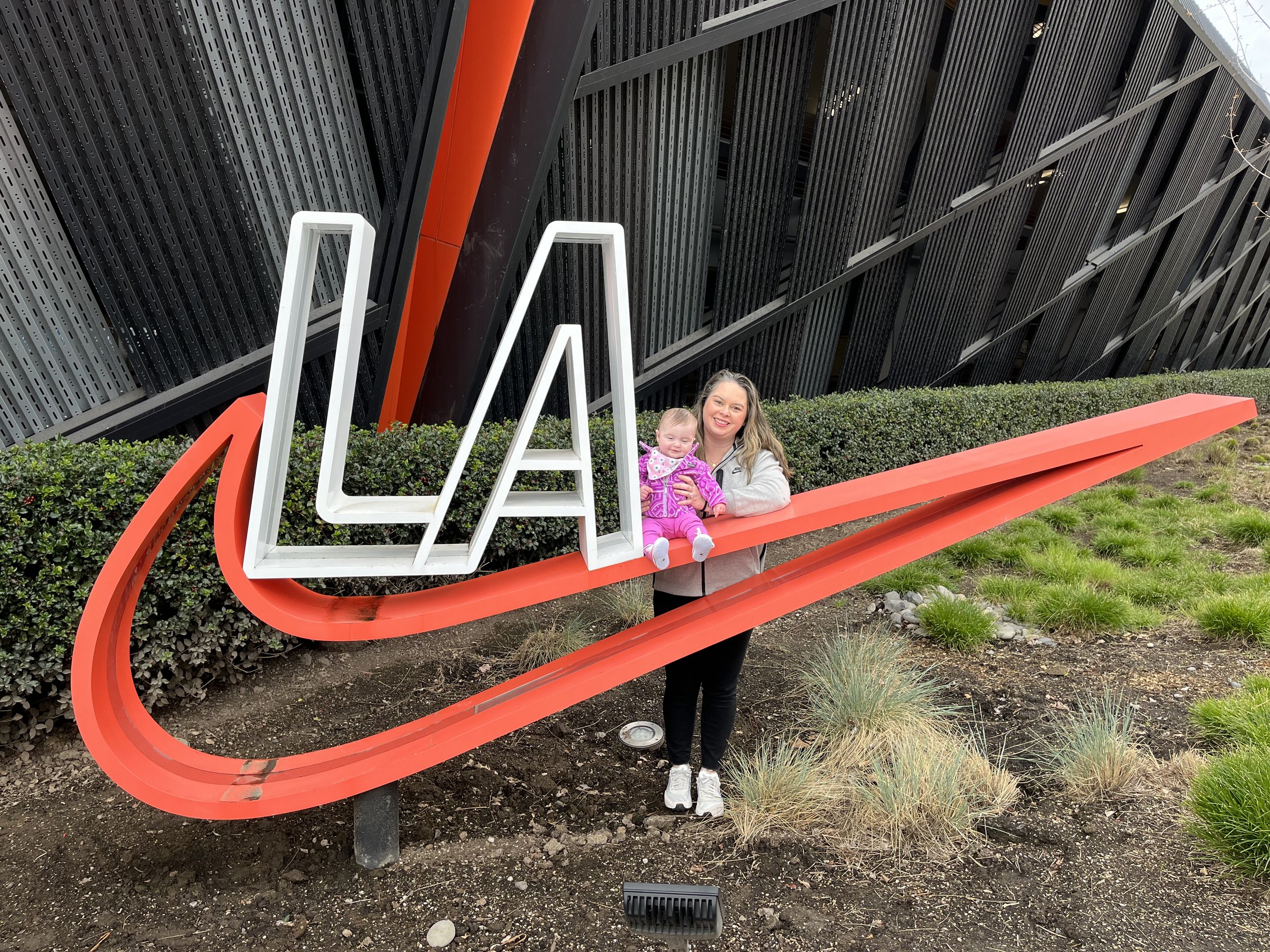 Woman holding a baby standing behind a large LA sign with a swoosh design, outdoors in front of a modern building with black and red accents.
