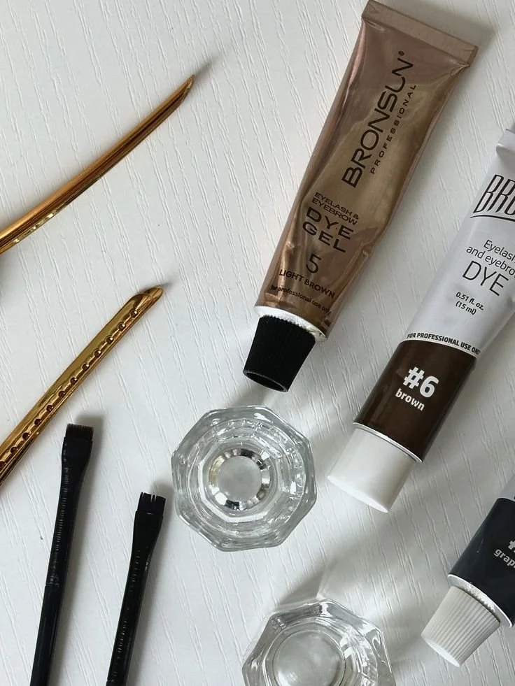Beauty tools and products including eyebrow and eyelash dye, brushes, and small glass bowls on a white surface.