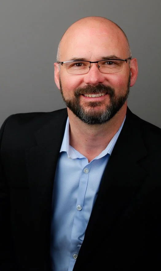 A professional headshot of a smiling, middle-aged man with a beard and glasses, wearing a light blue collared shirt and a black blazer, against a gray background.