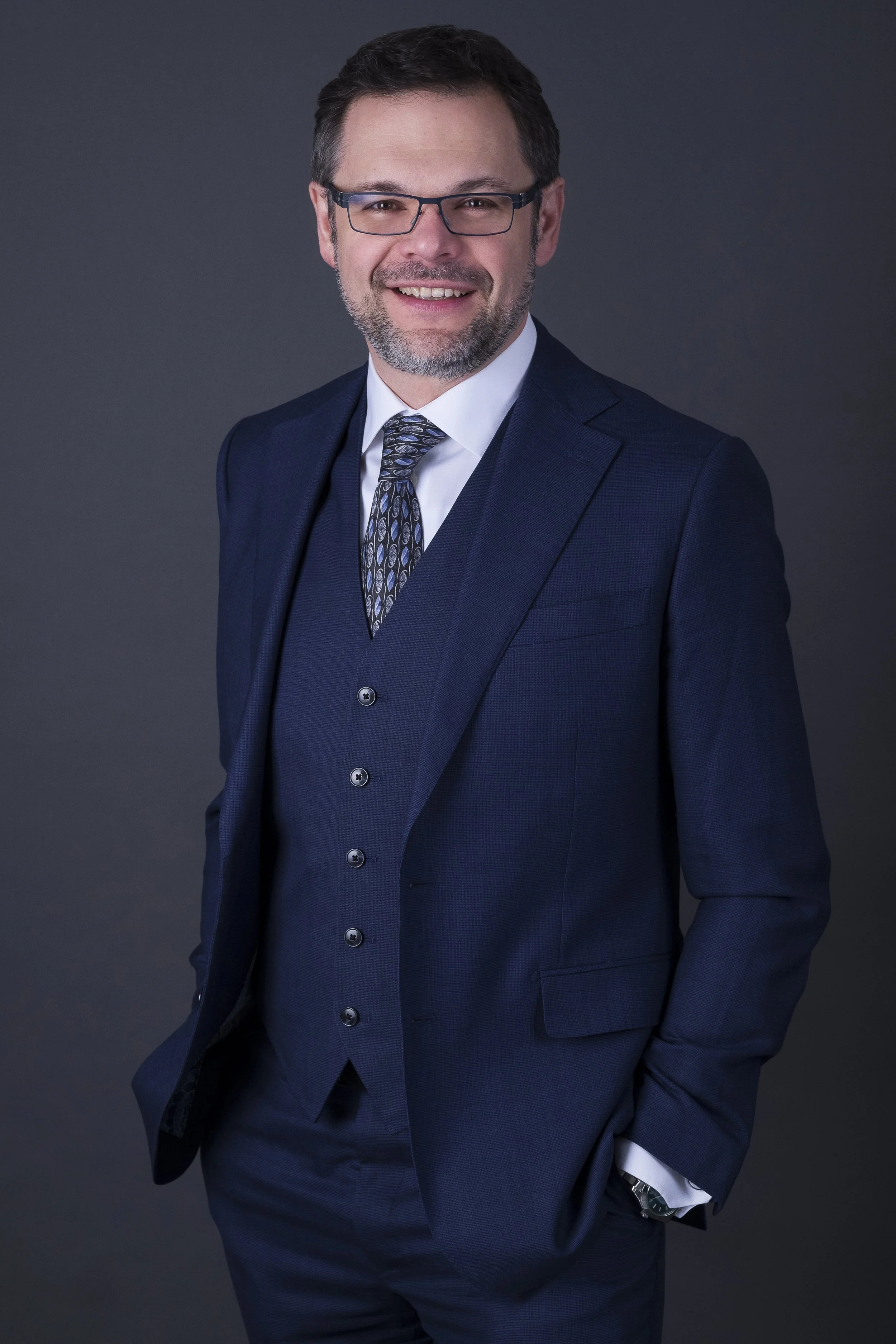 A man in a dark blue suit, white shirt, and patterned tie, smiling and looking at the camera, standing against a dark gray background.