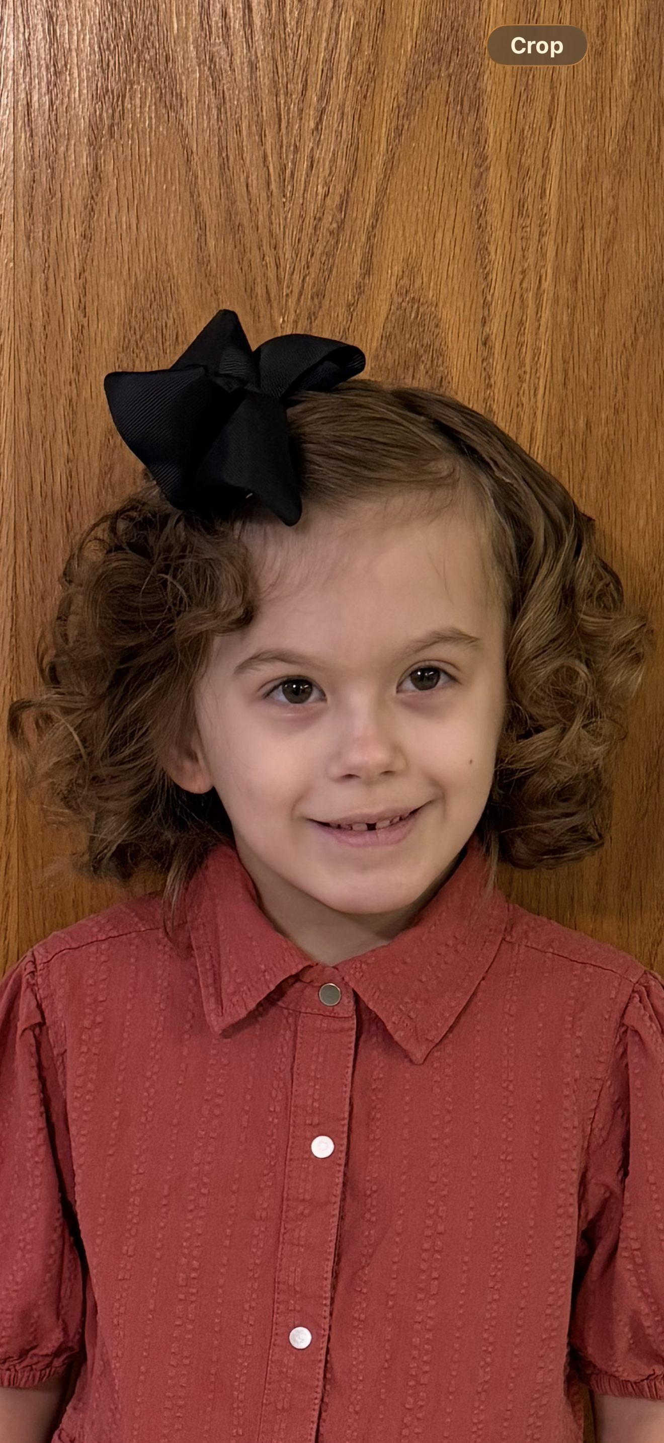 A young girl with curly brown hair wearing a large black bow and a red button-up shirt, standing in front of a wooden background.
