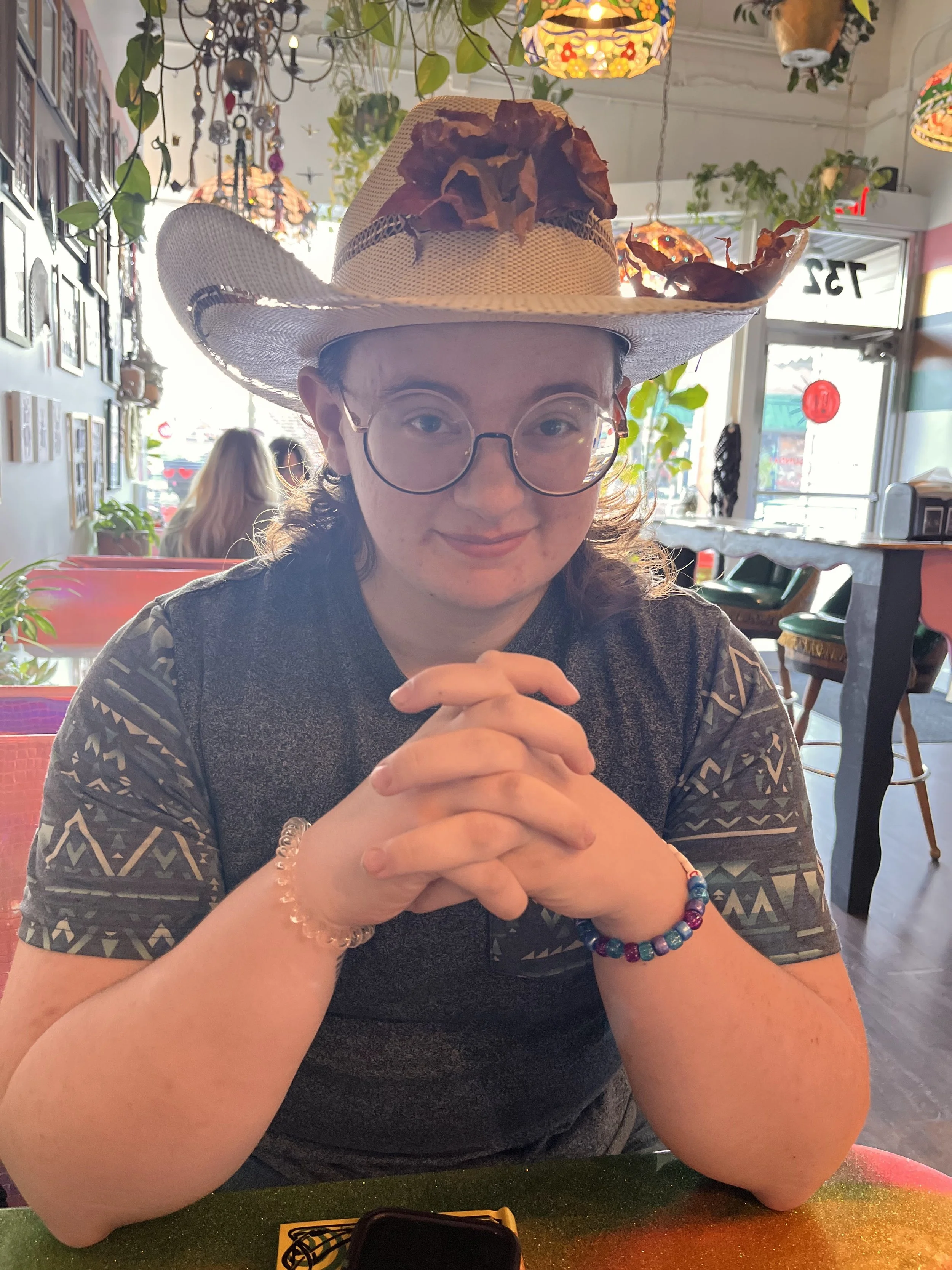 A woman sitting at a restaurant table, wearing glasses and a large fedora decorated with dried leaves and flowers, smiling with her hands clasped.