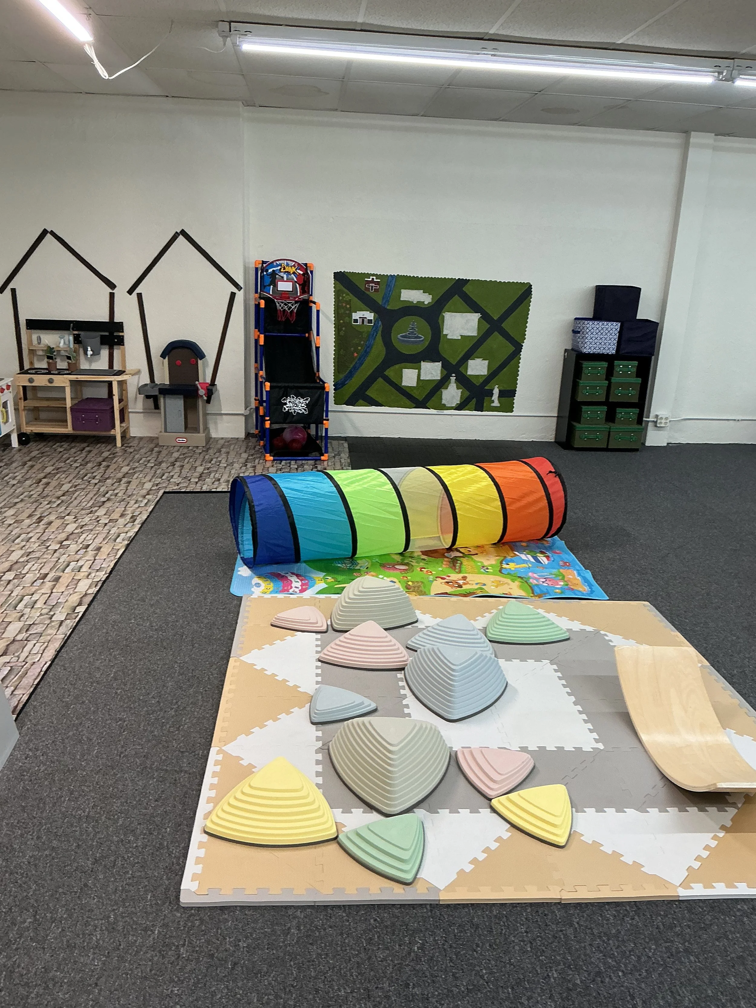 Indoor children's play area with colorful tunnel, foam stepping stones, and various toys including a basketball arcade game and a play mat with a colorful scene.