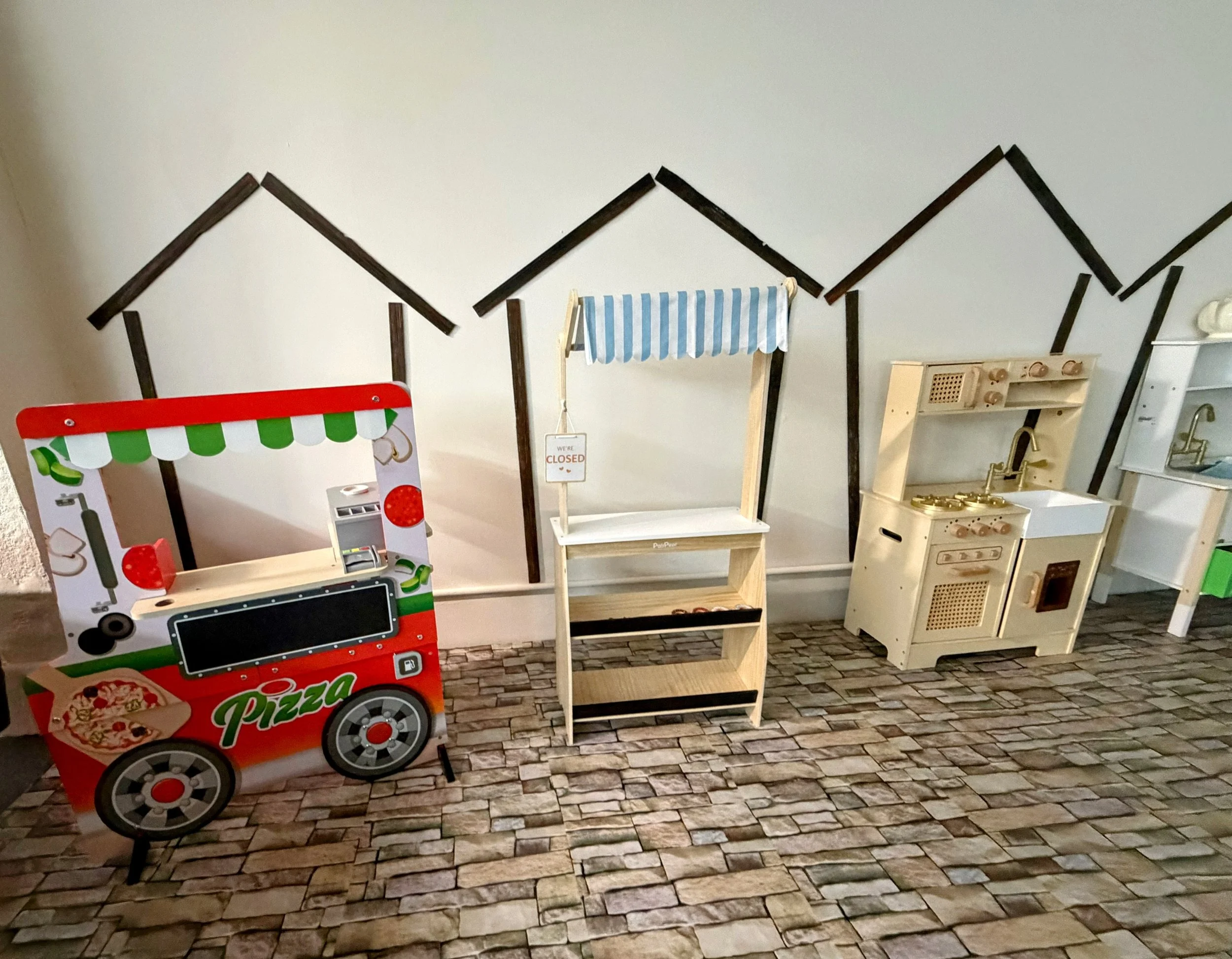 Collection of children's play kitchen toys and a pizza stand on a brick-patterned floor against a wall with black house outline decorations.