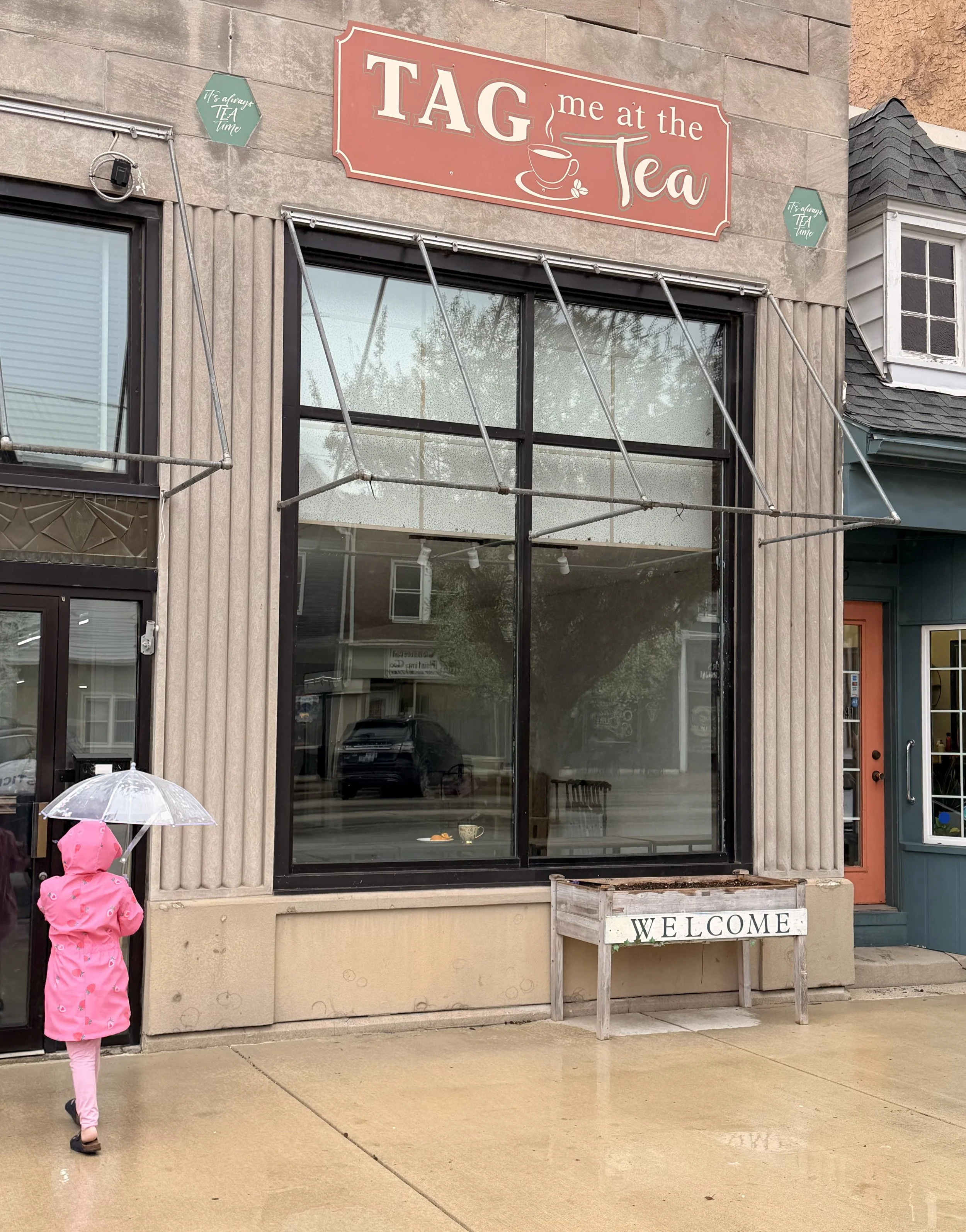  storefront with a red sign that reads 'TAG me at the Tea' and a large window. A girl in a pink raincoat is holding a transparent umbrella, standing on a wet sidewalk outside the store.