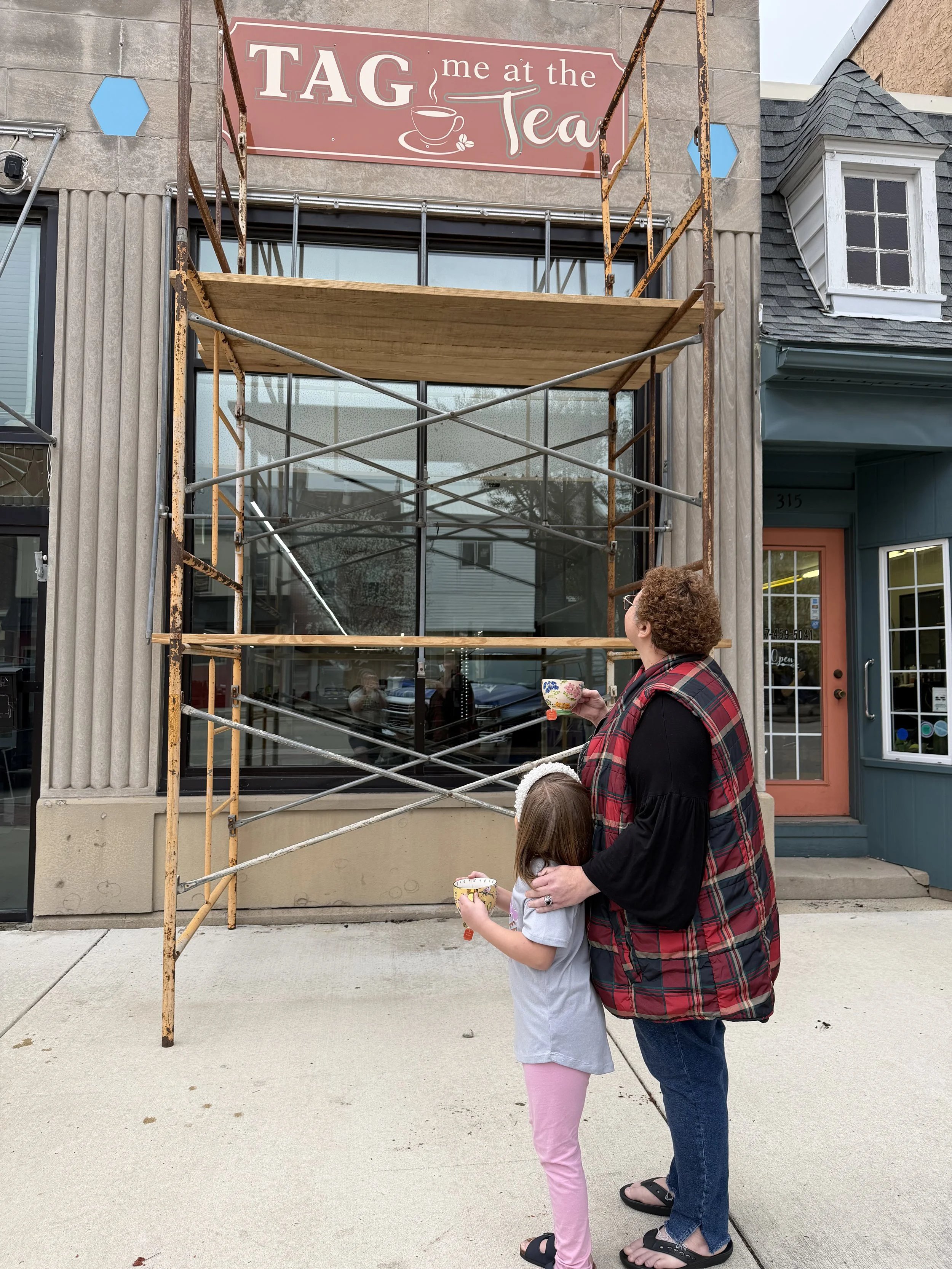 A woman and a young girl standing outside a storefront with a sign that reads 'TAG me at the Tea.' The woman is holding a bowl and the girl is also holding a bowl, both seem to be enjoying tea. There is a scaffolding structure in front of the window.