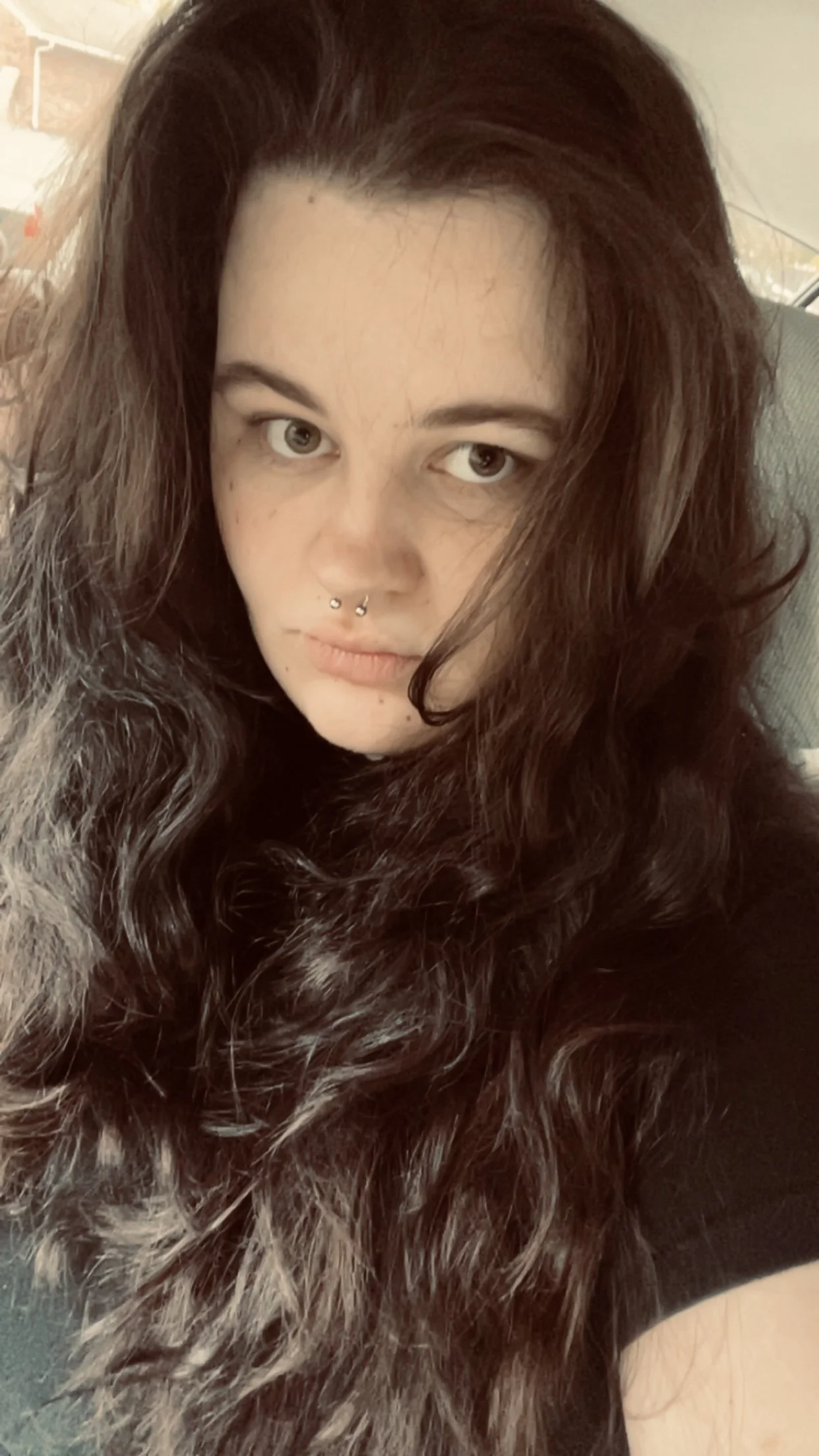 Close-up of a young woman with long, wavy dark hair and light skin, looking into the camera, with a septum piercing, in a casual setting
