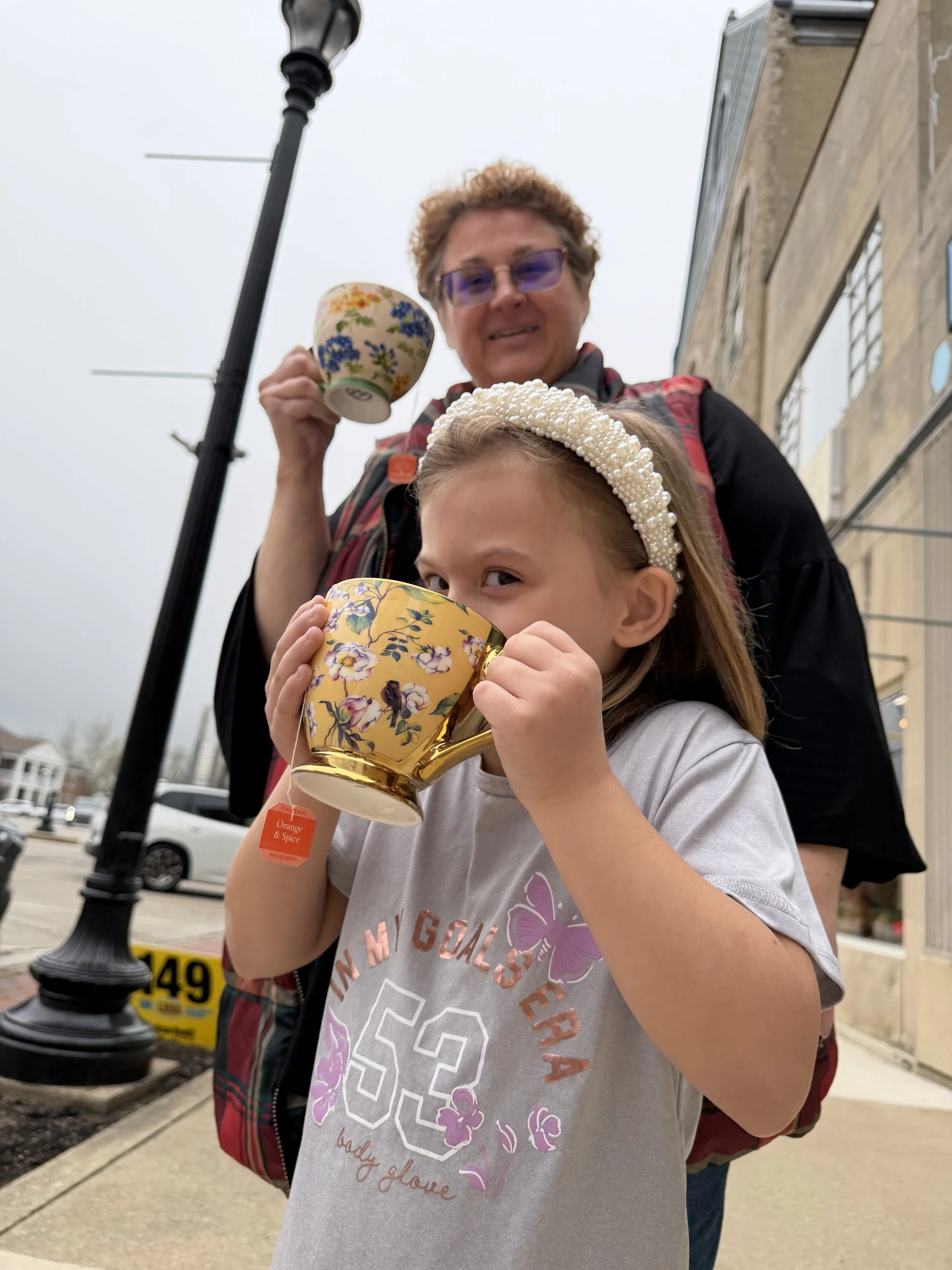 A young girl and an older woman drinking tea outside. The girl is holding a large floral mug and the woman is holding a smaller floral mug, both smiling and looking at the camera. It's a cloudy day, and they are standing on a sidewalk near a storefront.