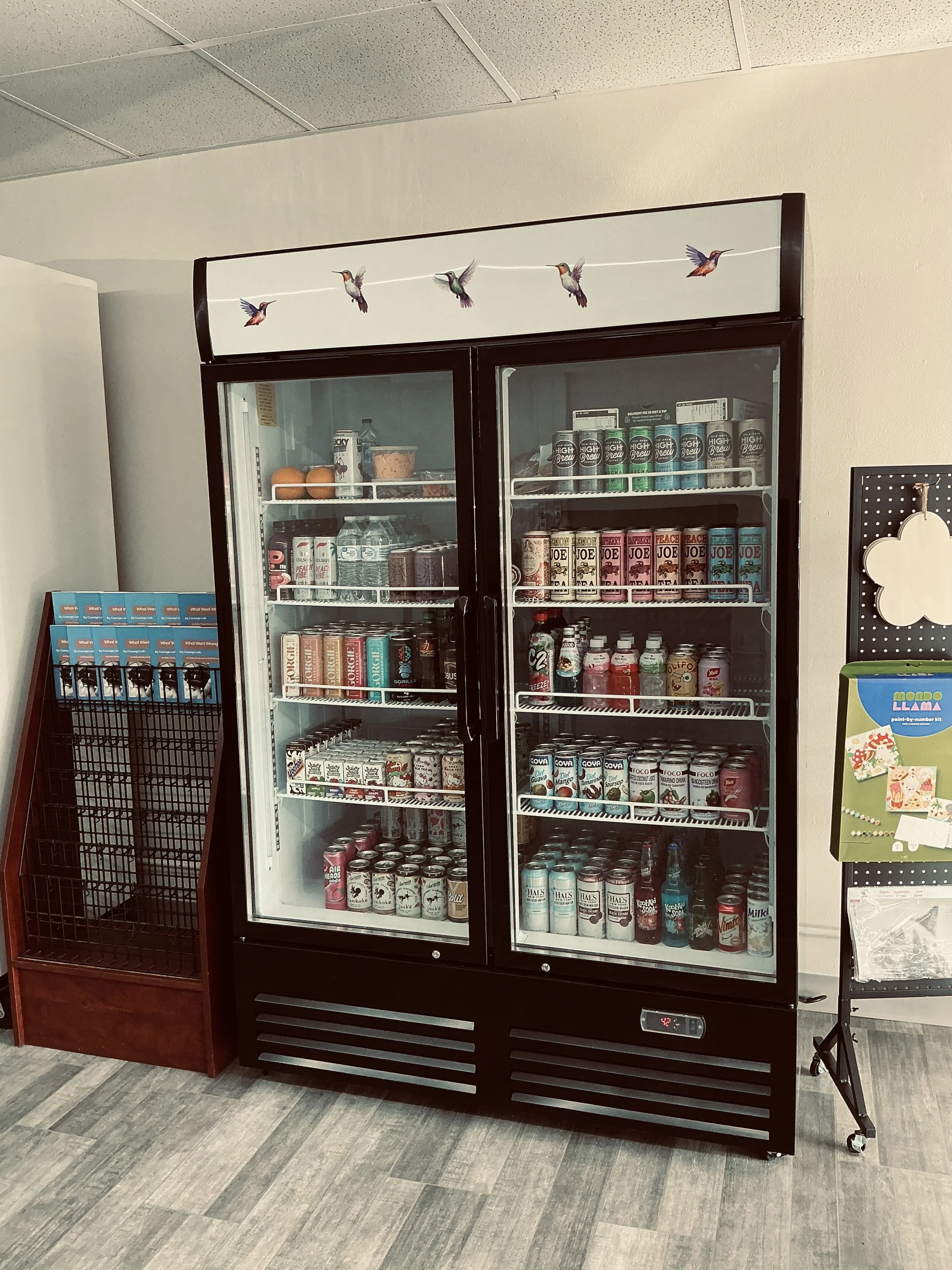 A black refrigerated display case filled with various canned drinks and beverages, standing next to a wire rack with blue packets and an adjacent pegboard with decorative items.