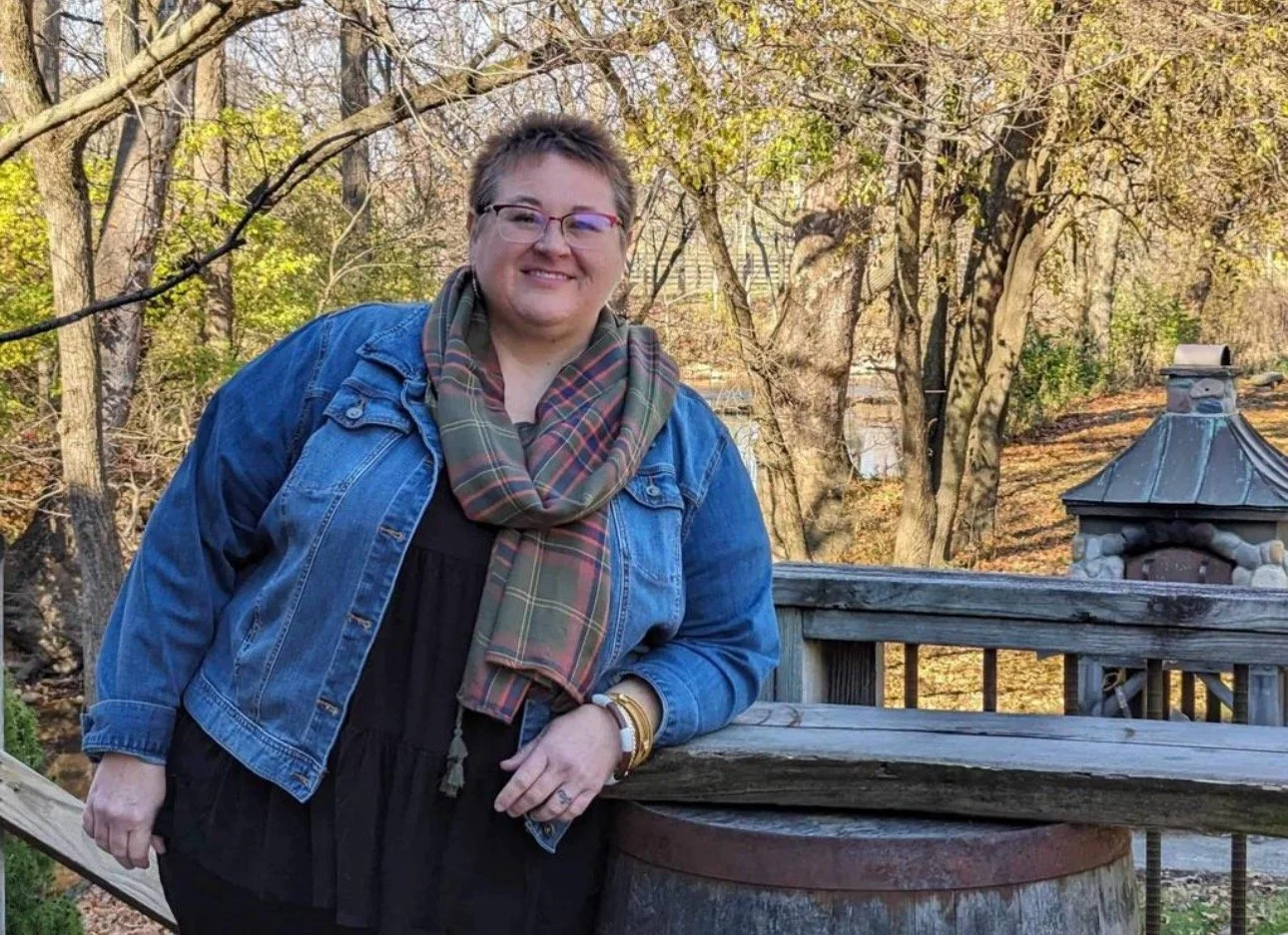 A smiling woman leaning on a weathered wooden railing outdoors in a wooded area with fall foliage.