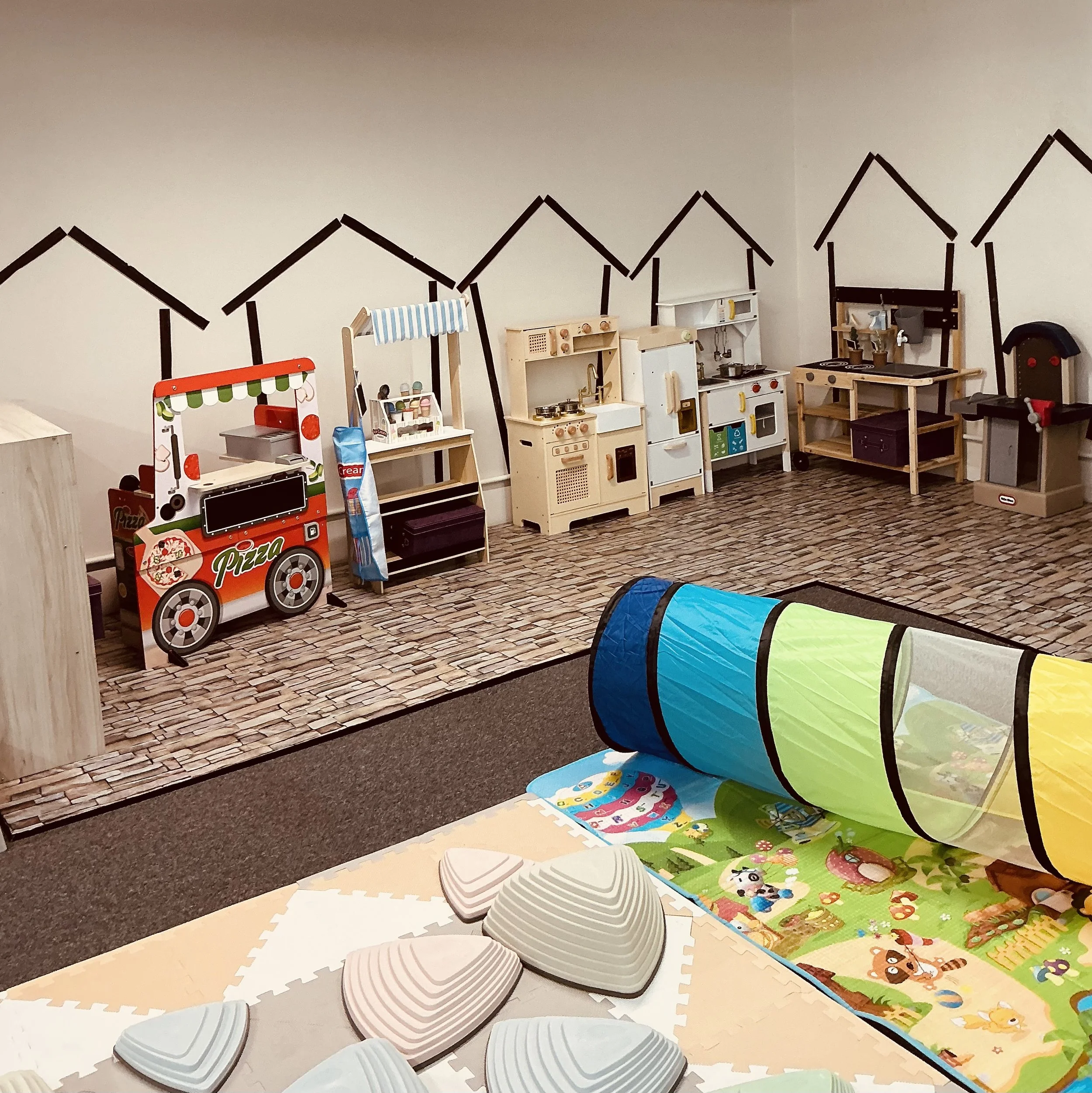 Children's play area with toy kitchens, a pizza cart, and a playhouse tunnel on a colorful rug inside a room with a wall decorated with house outline mural
