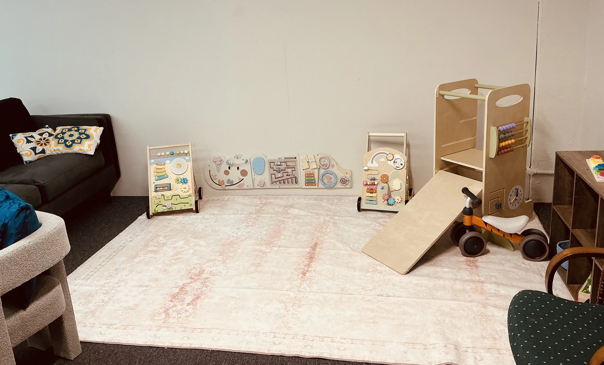 Indoor play area with wooden activity toys, a small slide, and a black couch with colorful pillows, on a beige rug.