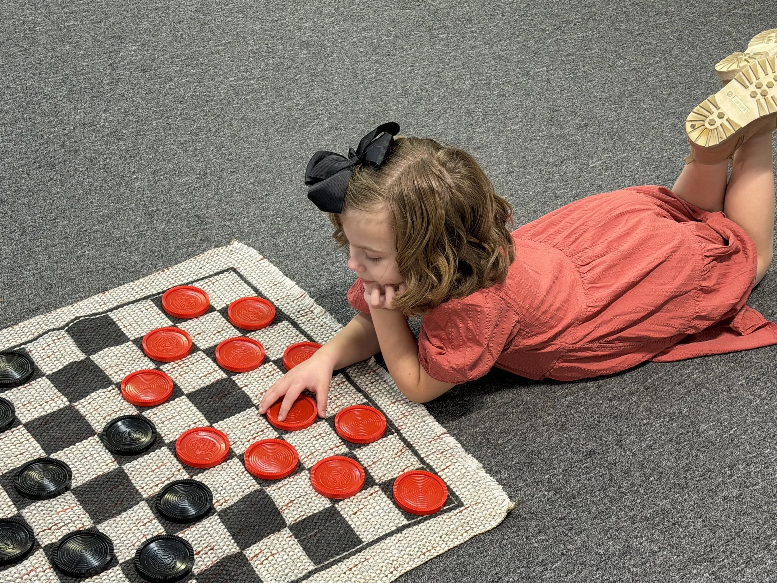 A young girl with a black bow in her hair, lying on her stomach on a gray carpet, playing checkers with red and black circular pieces on a woven board.