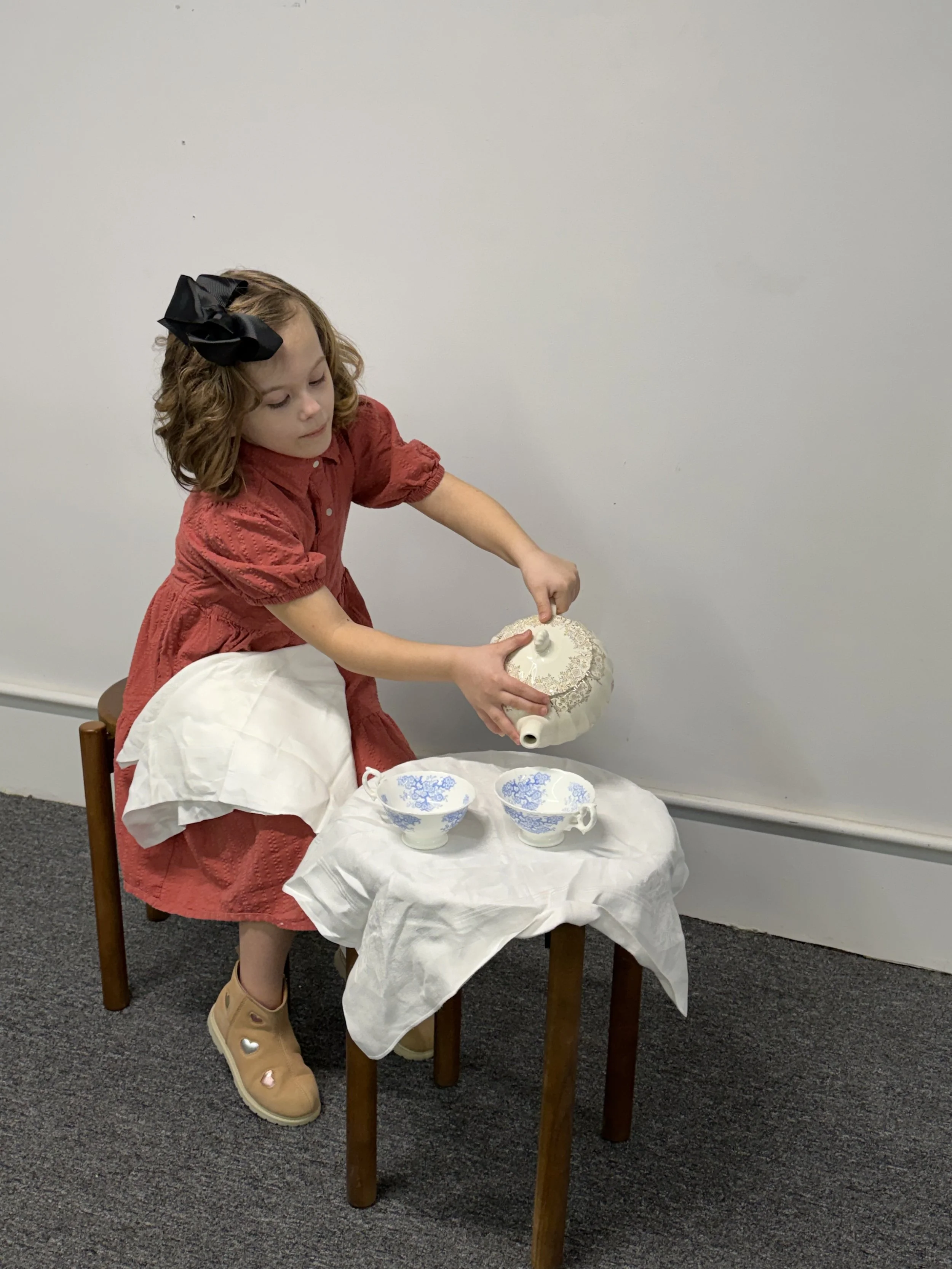 A young girl with curly hair wearing a red dress and a large black bow in her hair is pouring tea from a teapot into two blue-patterned bowls on a small table covered with a white cloth.