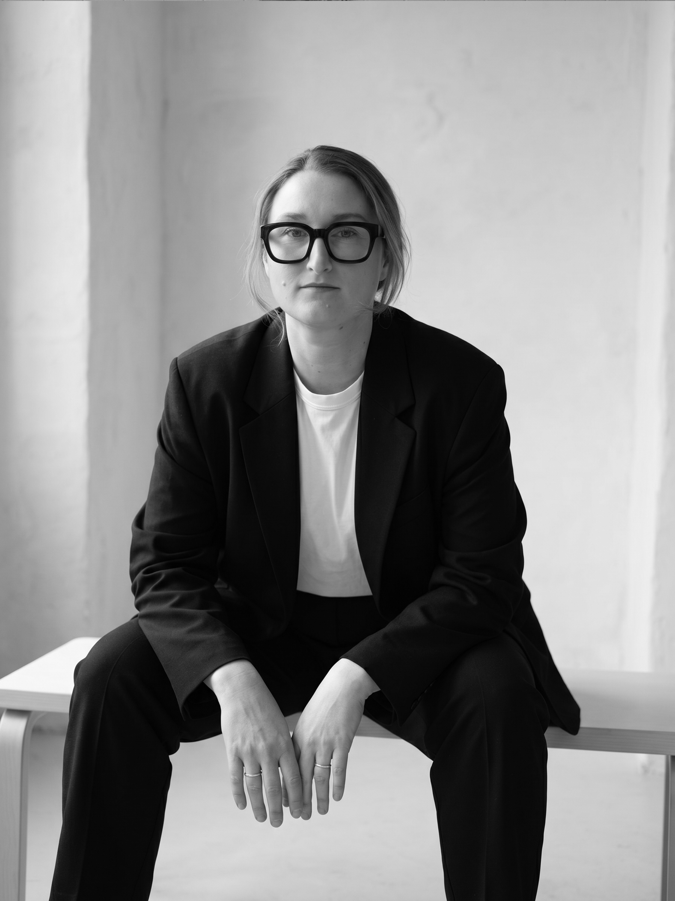 A woman with short hair, large glasses, wearing a black blazer over a white top, sitting on a bench against a plain wall. She is looking directly at the camera with a neutral expression.